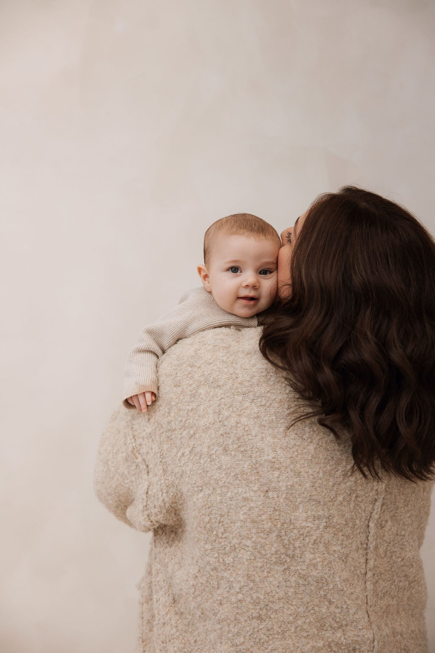 Tender moment of mother kissing infant on cheek, baby peeking over mothers shoulder in matching cream outfits.