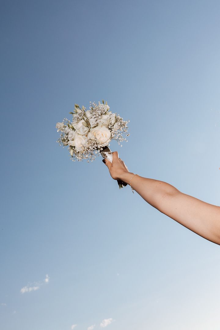 A hand holding a white bridal bouquet of roses and baby's breath raised against a clear blue sky.