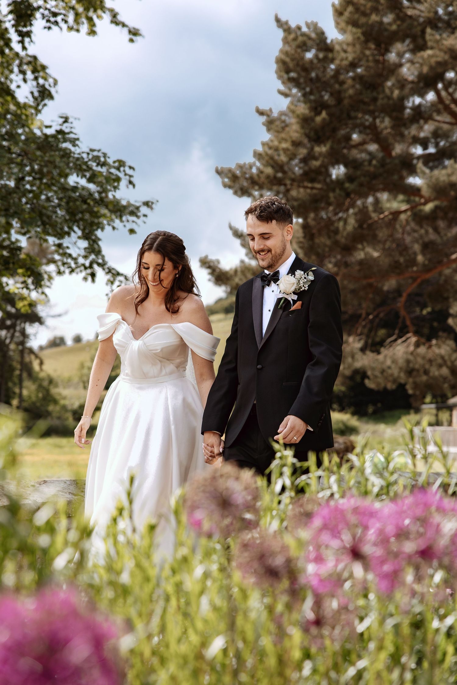 Smiling bride and groom walk hand in hand through a garden with colorful wildflowers.
