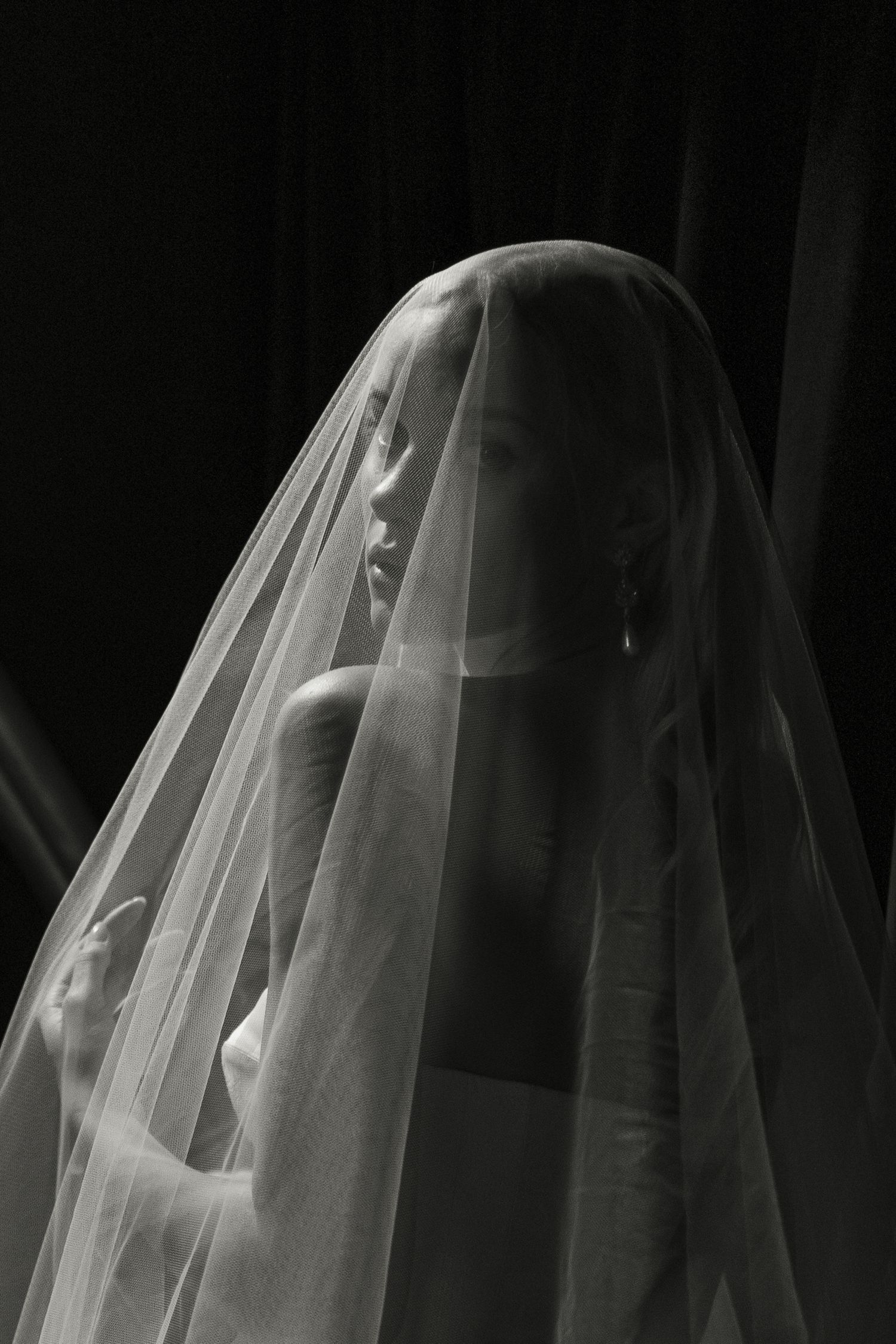Black and white portrait of a woman draped in a sheer white veil against a dark background.