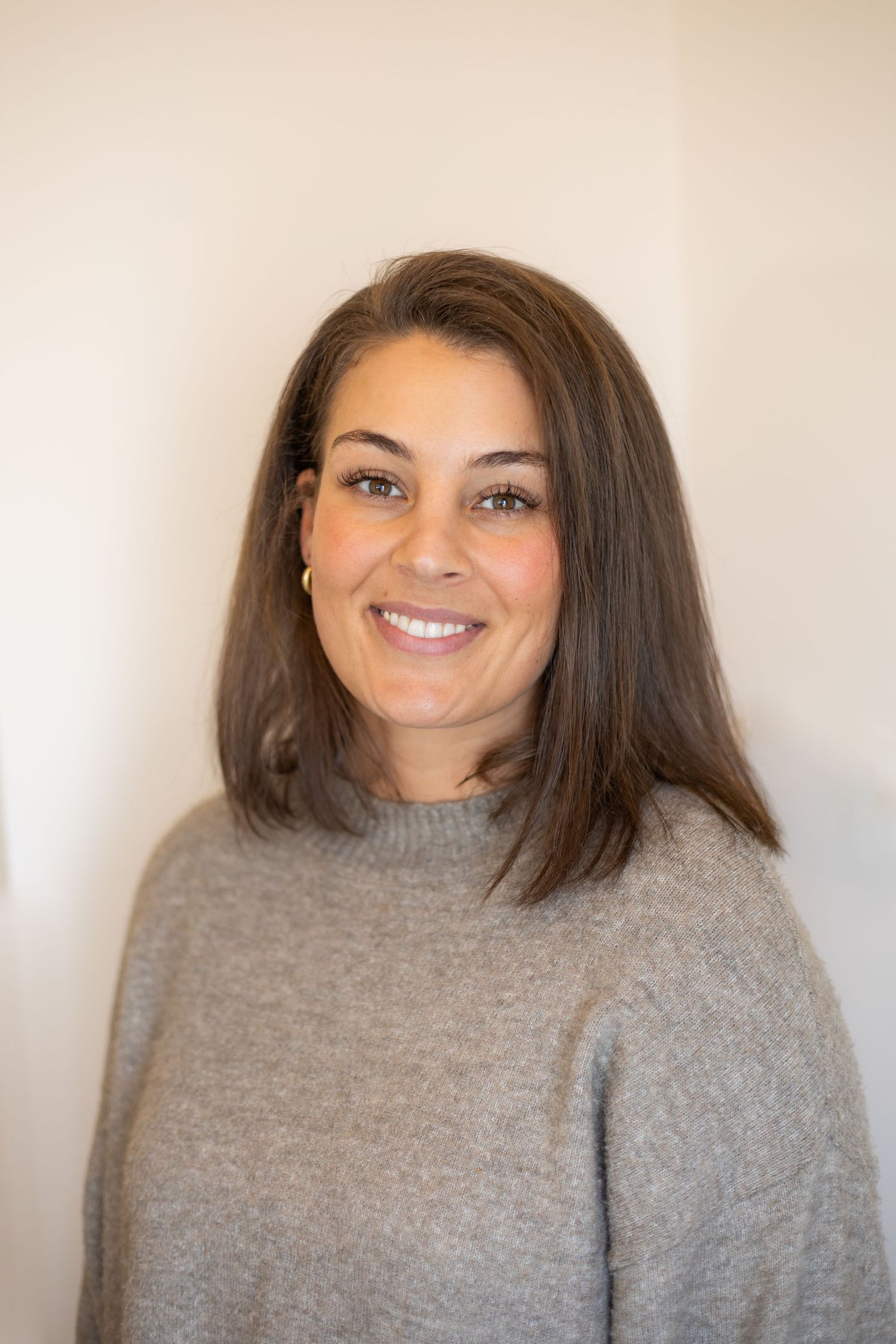 Smiling brunette woman with shoulder-length hair wearing a gray turtleneck sweater against a white background.