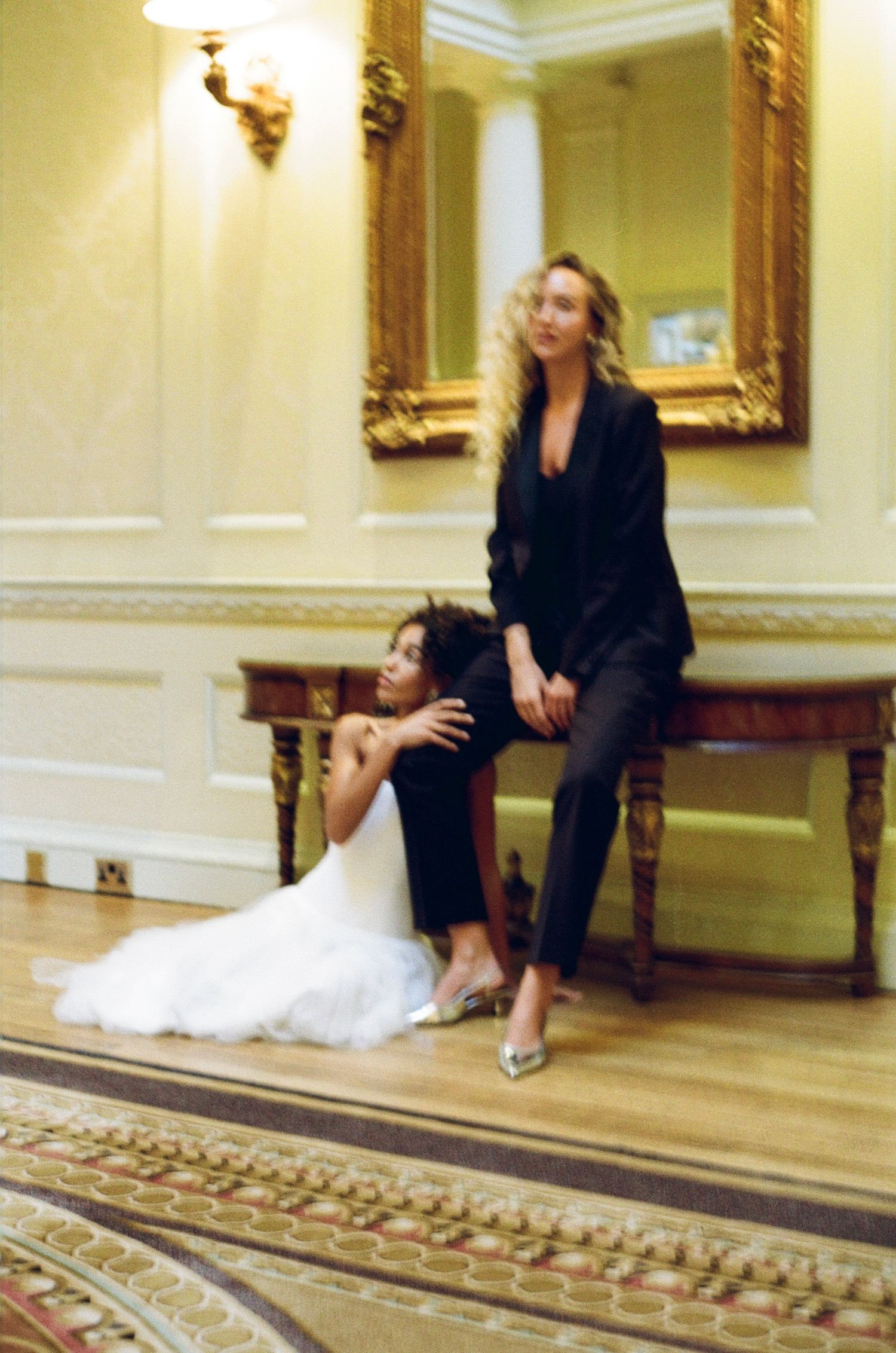 Two women in elegant attire pose in a grand hotel lobby with gold-framed mirror, one in white tulle gown.