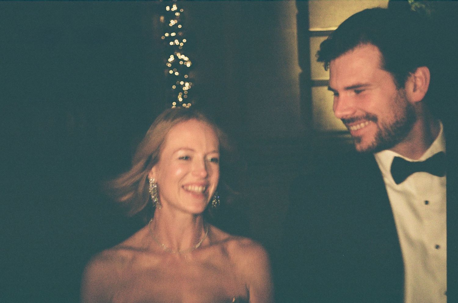 Bride in strapless gown and groom in tuxedo share a joyful moment under warm fairy lights at reception.