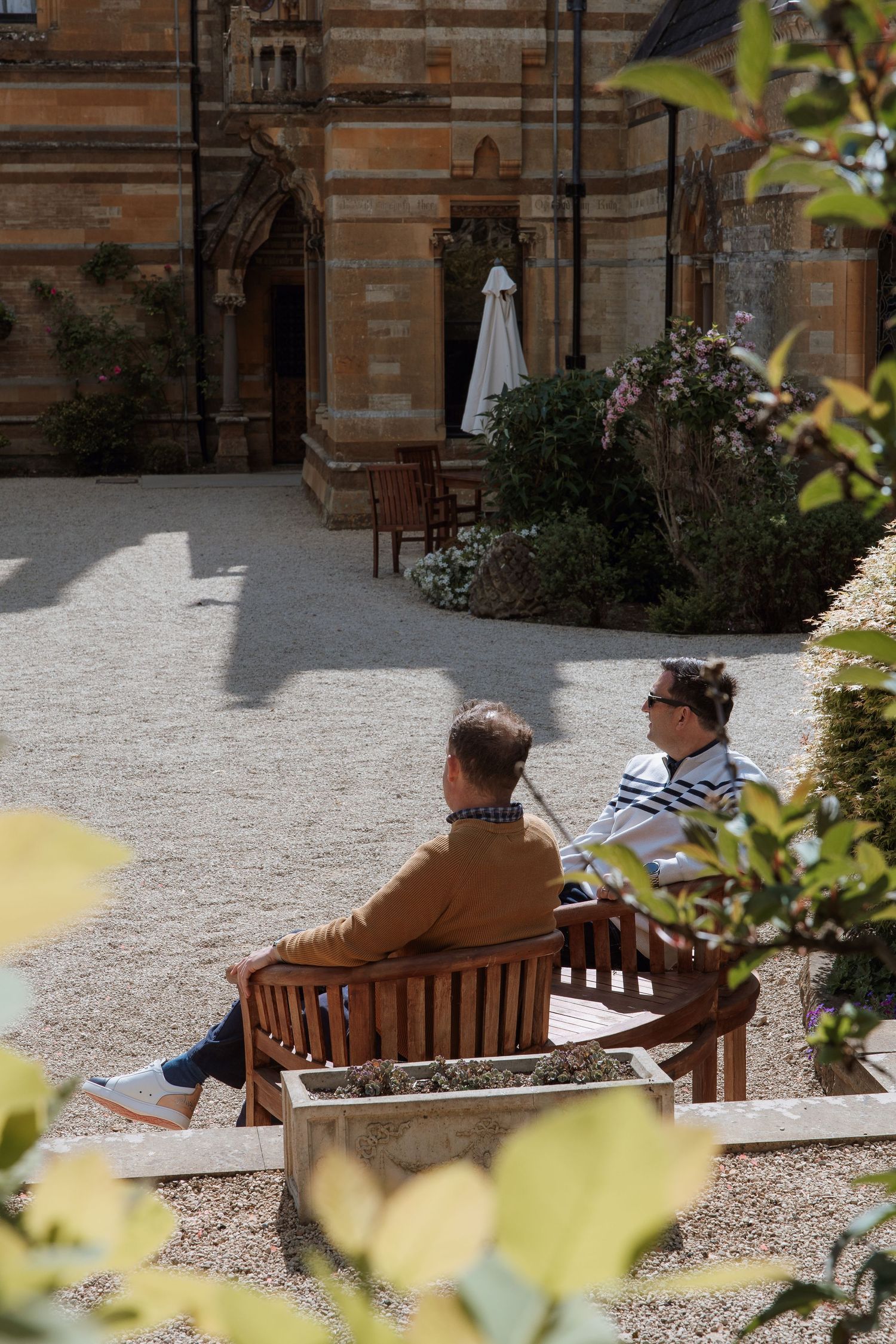 Two people sitting on a wooden bench in a sunlit courtyard garden beside a historic stone building.