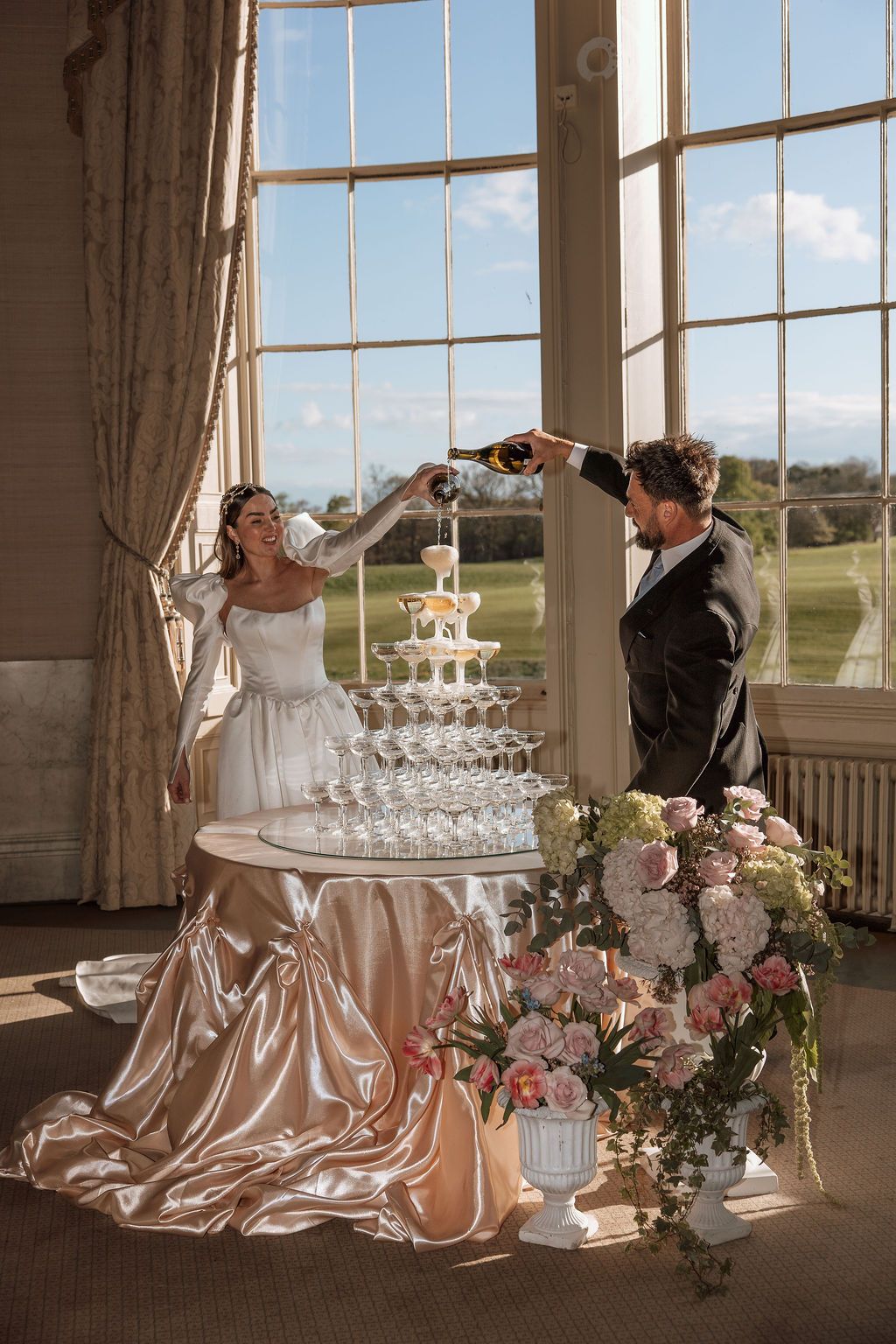 Bride and groom pouring champagne tower at elegant wedding reception with floral arrangements and large windows. York wedding photographer
