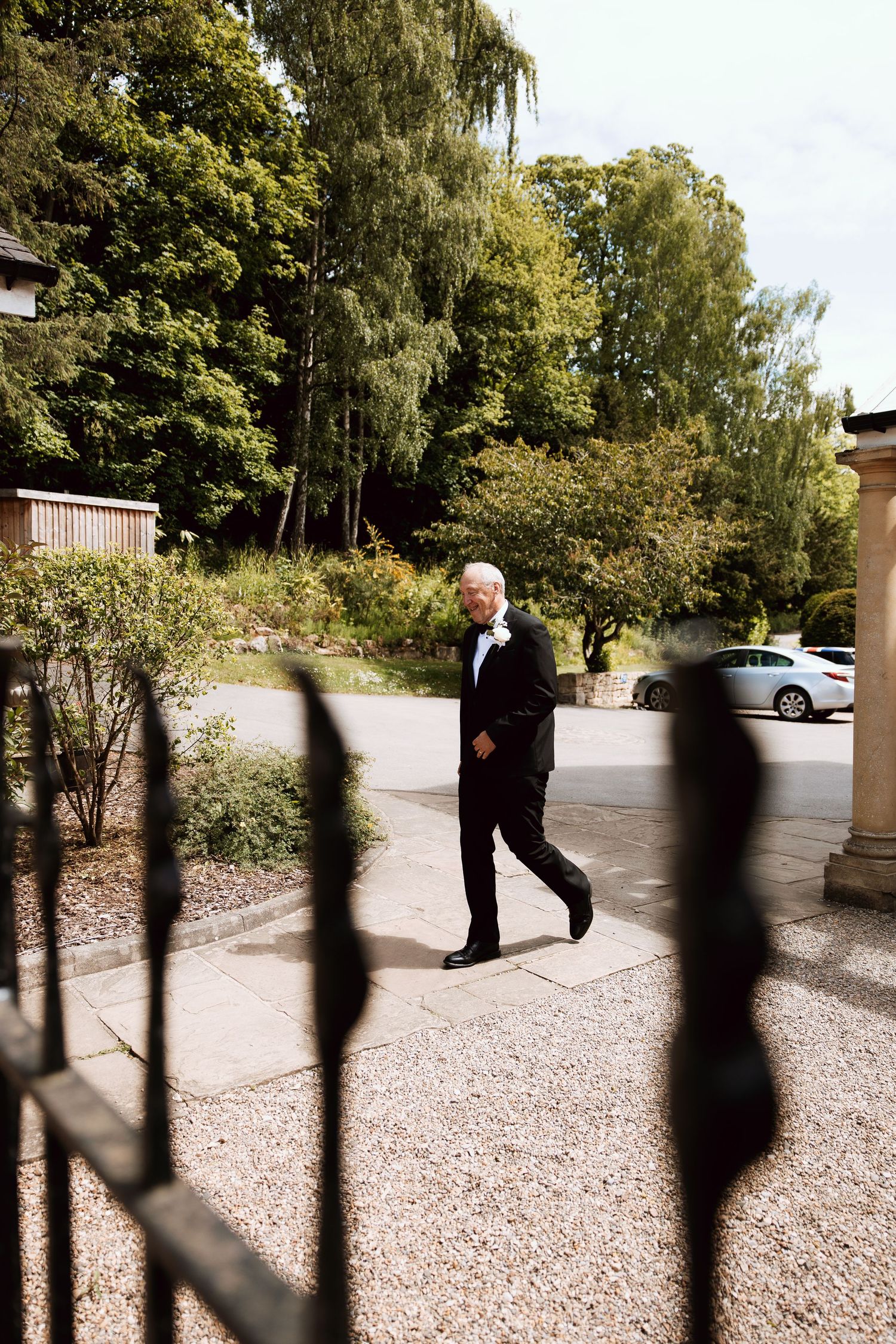 Elegant older gentleman in black tuxedo walking through ornate iron gates at a garden wedding venue.