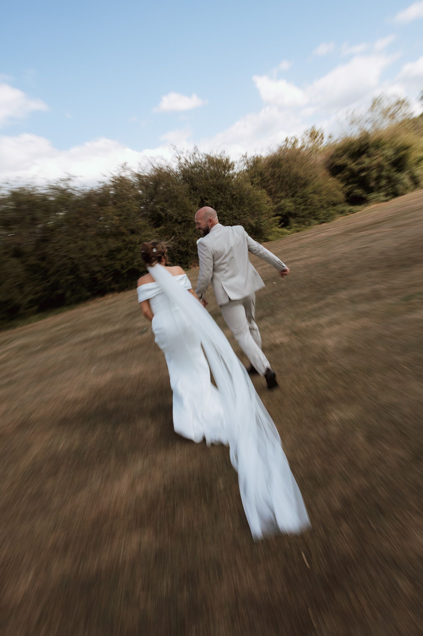 Bride and groom running through a field on their wedding day, motion blur capturing joyful movement.