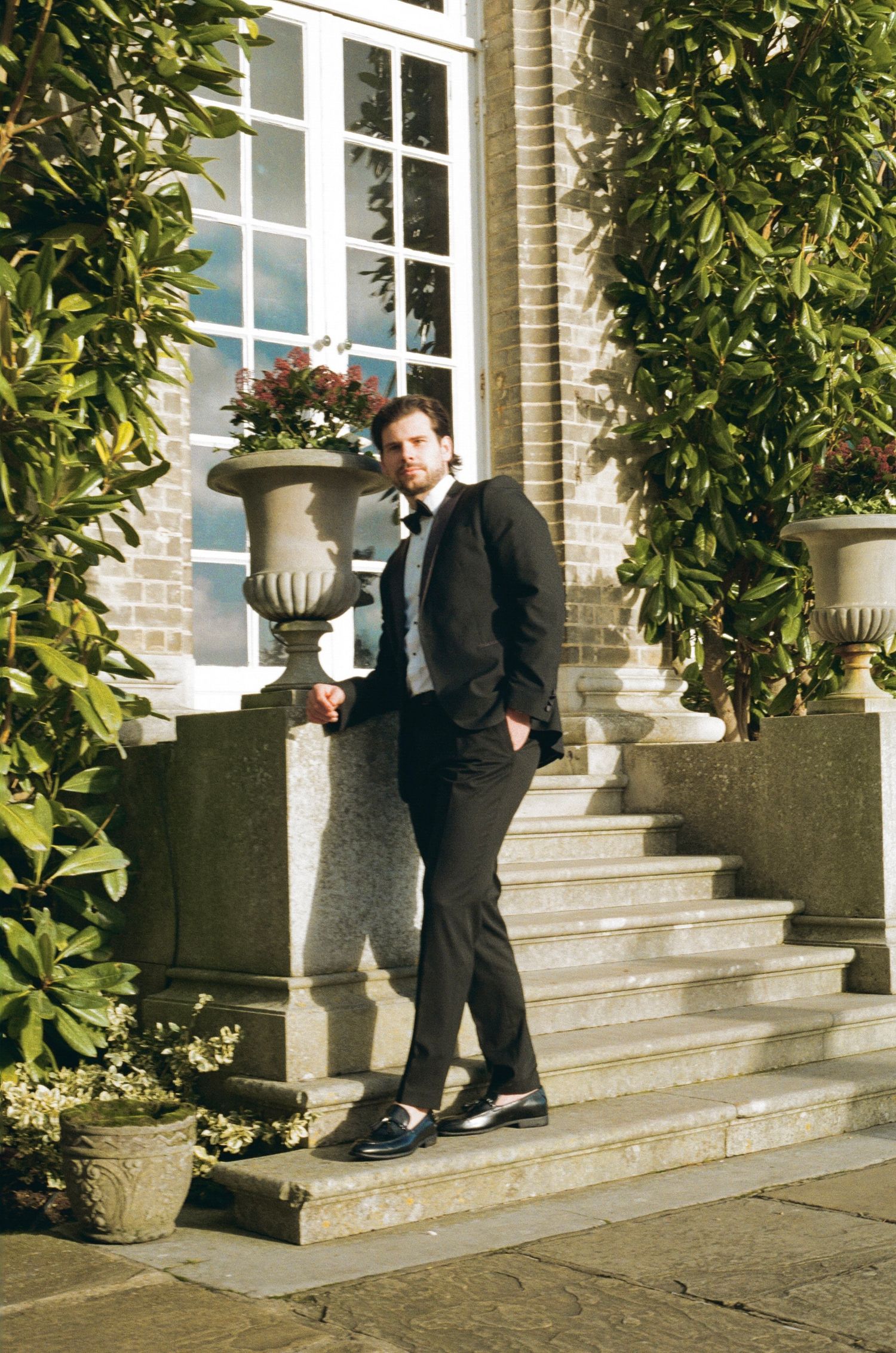 Dapper groom in black suit leaning against stone steps with urn planters at a classic estate wedding.