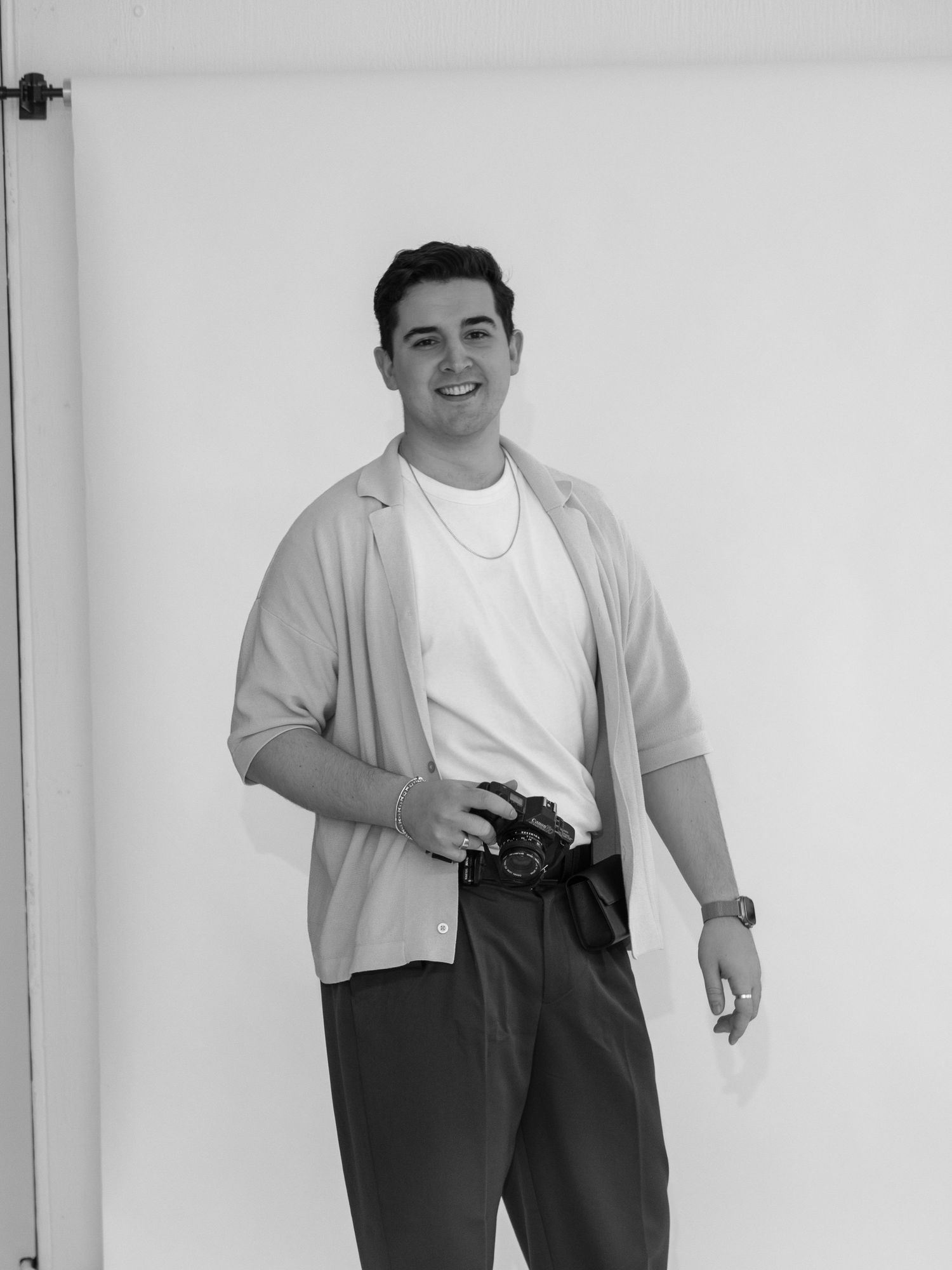Smiling young man holding a camera wearing a light cardigan and white tee in a black and white photo.