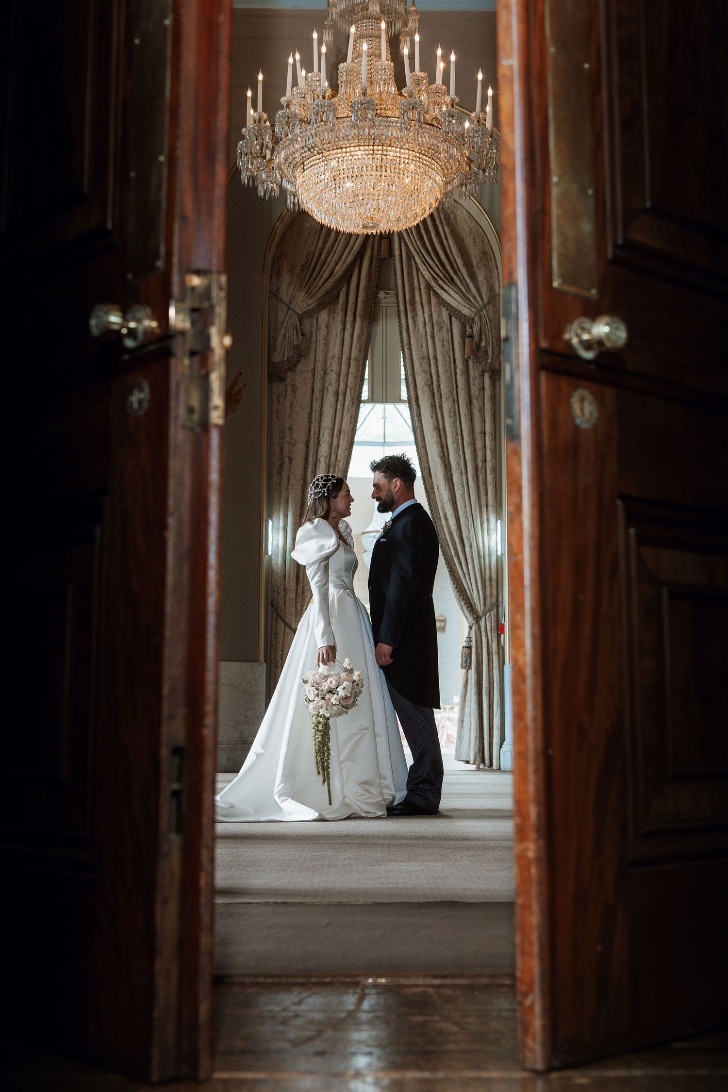 Elegant bride and groom stand beneath crystal chandelier in grand hall, viewed through ornate wooden doors.