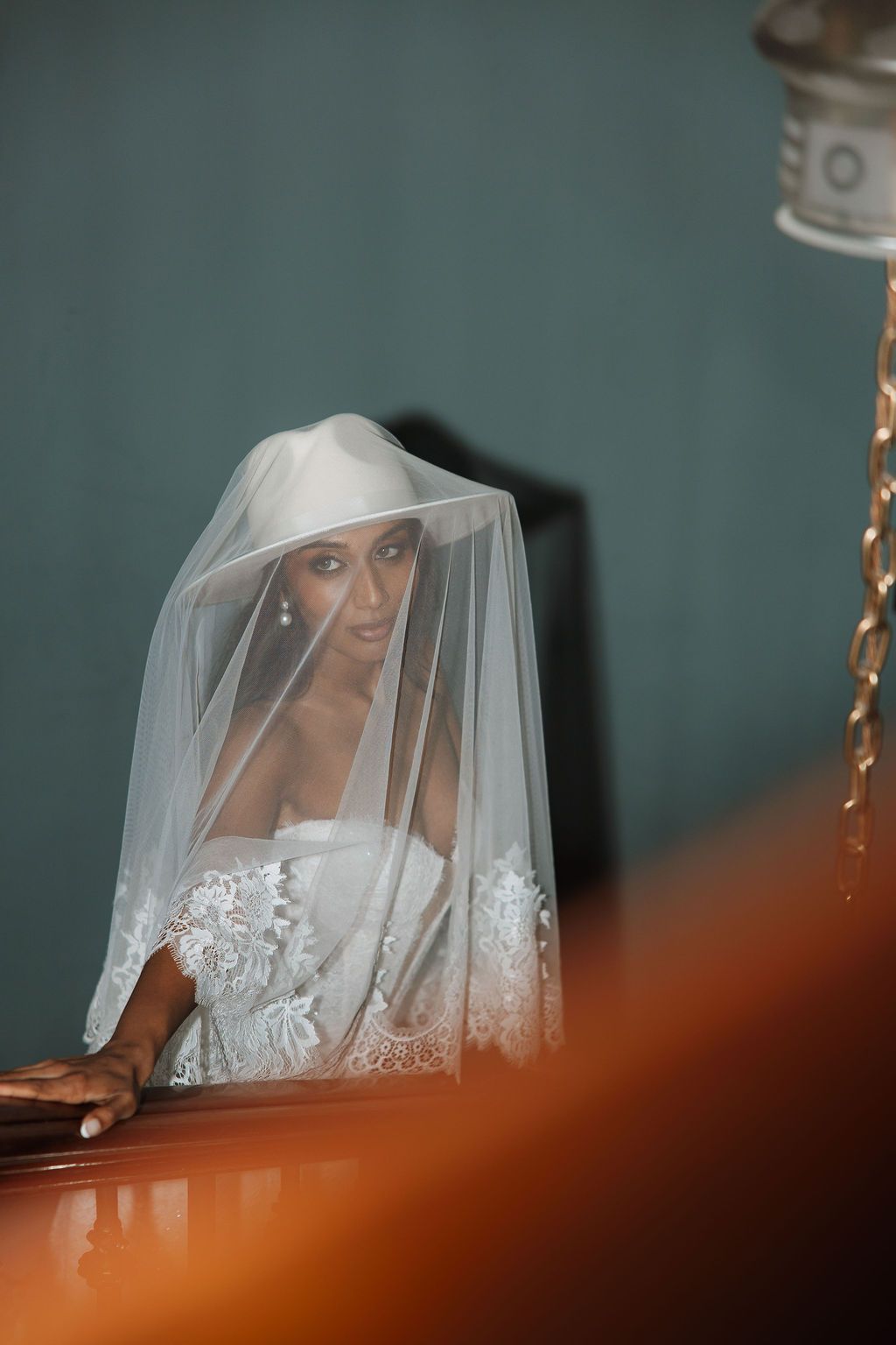 Bride in white lace wedding dress and veil draped over head, seated in church pew with soft ambient lighting.