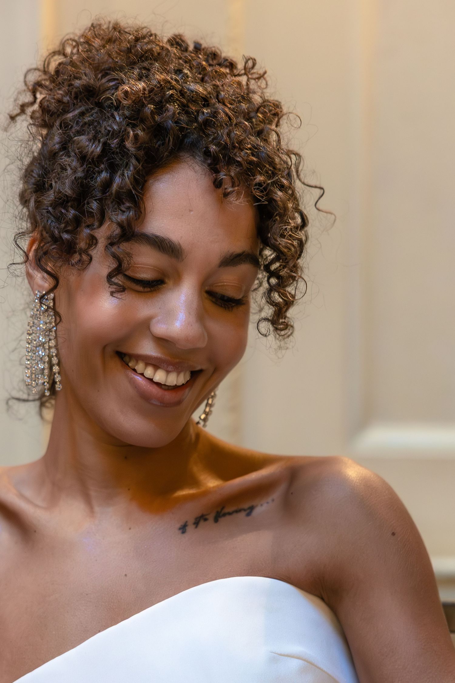 Smiling bride with curly hair and chandelier earrings wearing a white strapless dress indoors.