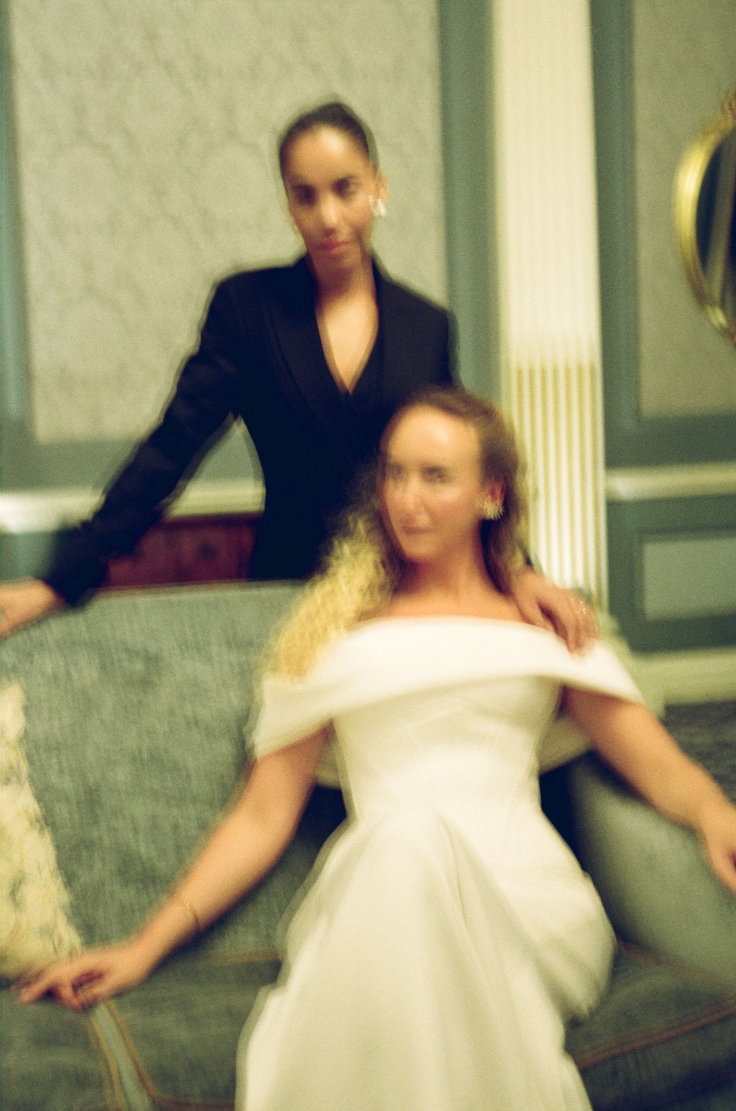 Two women pose elegantly in a blurred film photo, one in white off-shoulder gown seated on vintage sofa, other in black blazer.