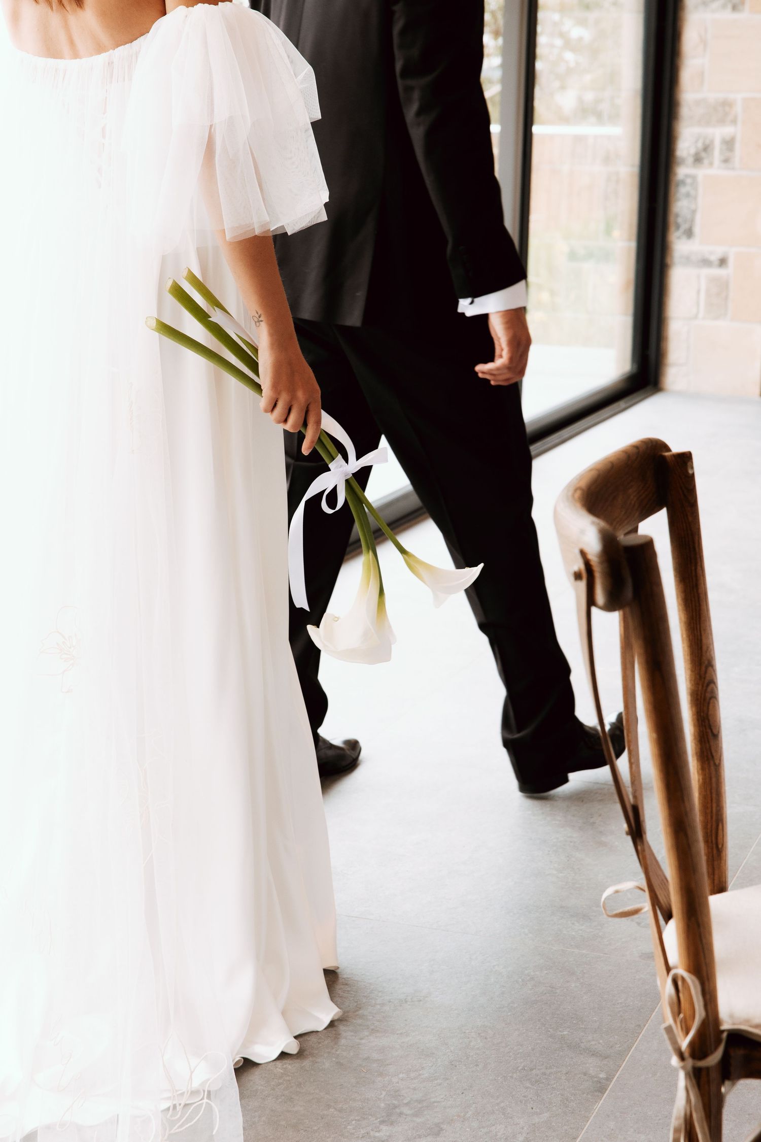 Bride in white flutter-sleeve gown holds calla lily bouquet beside groom in black suit at ceremony.