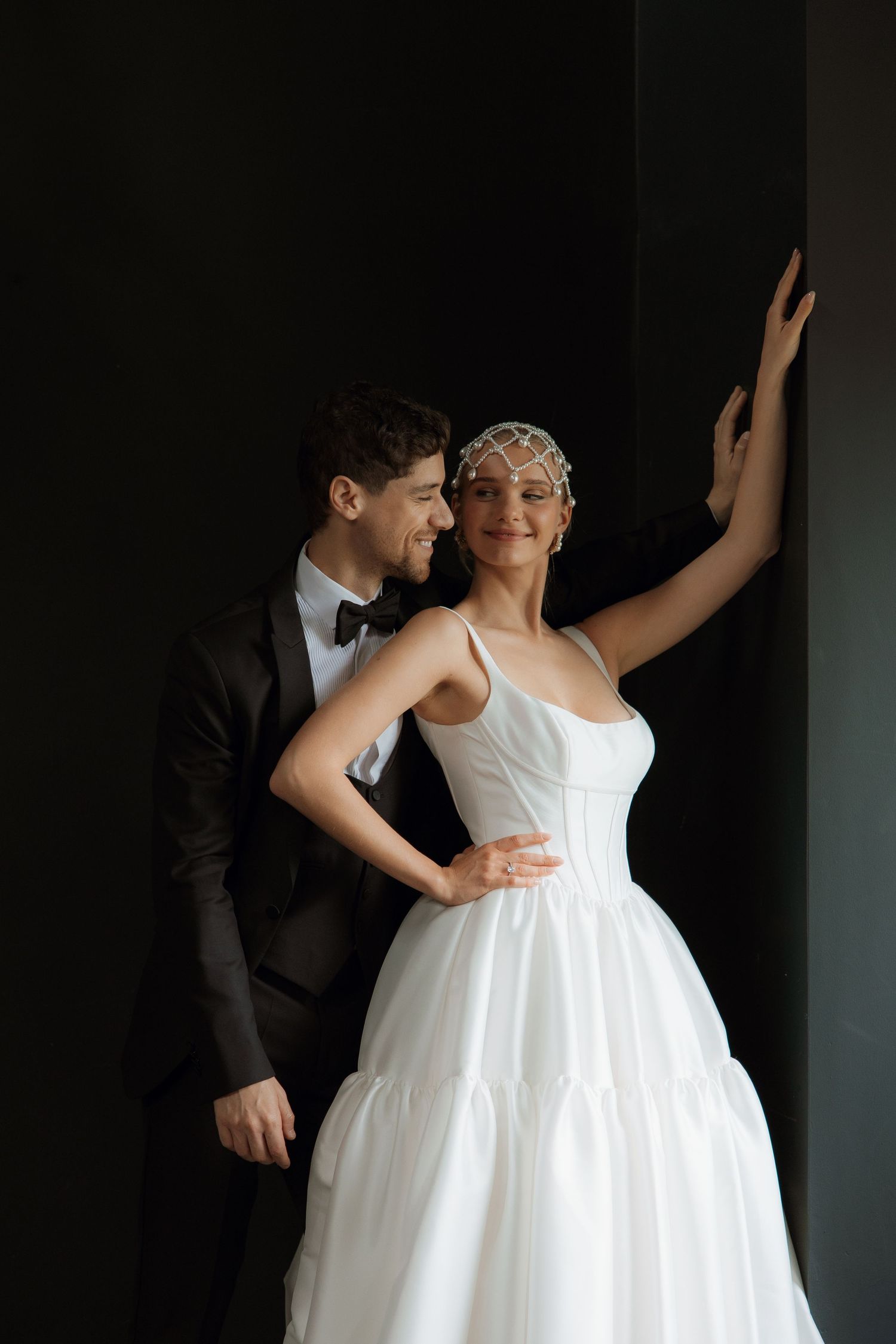 A bride in a white ballgown and pearl headpiece poses with a groom in a black tuxedo against a dark wall.