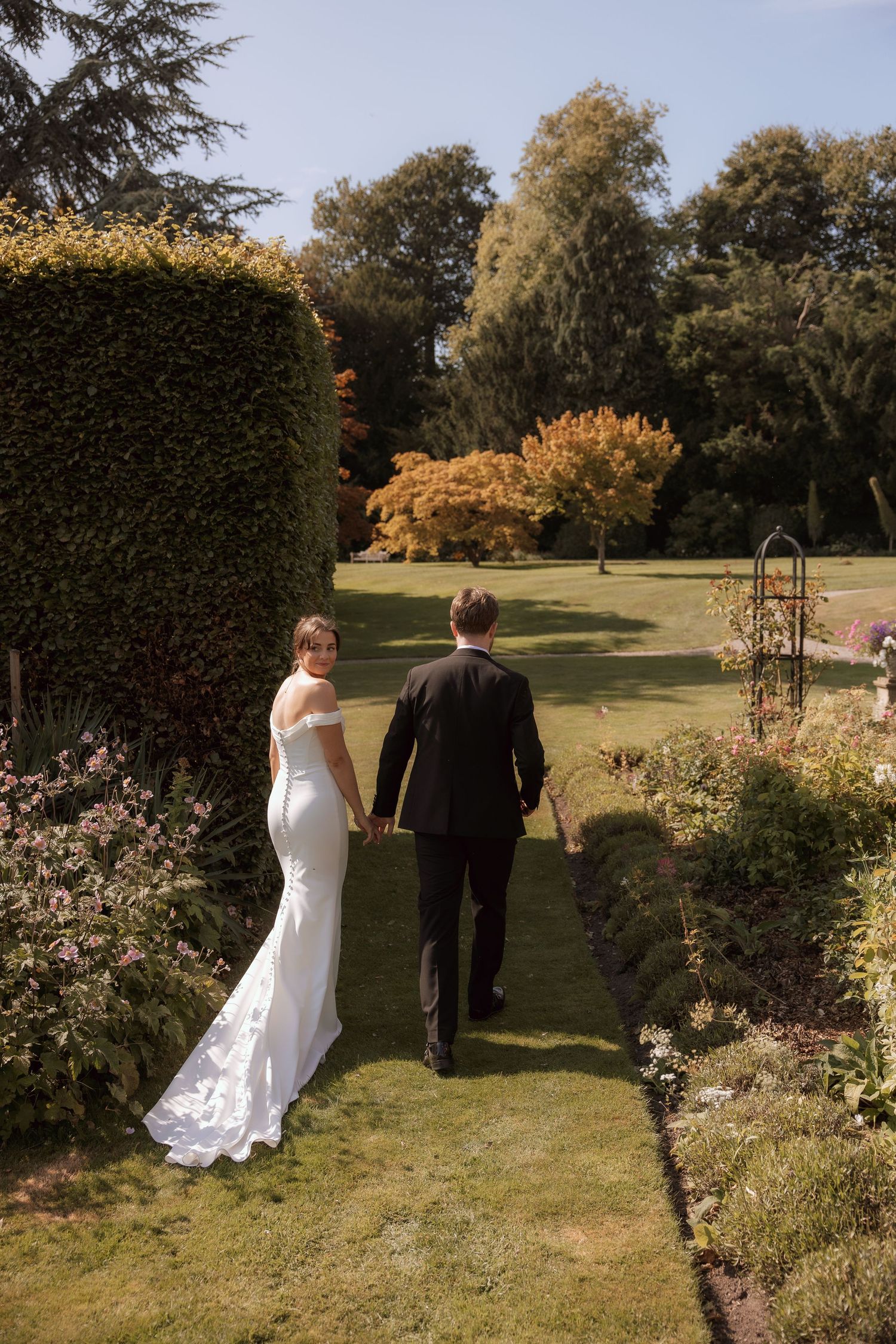 Bride and groom walking hand in hand through a formal garden pathway on their wedding day.