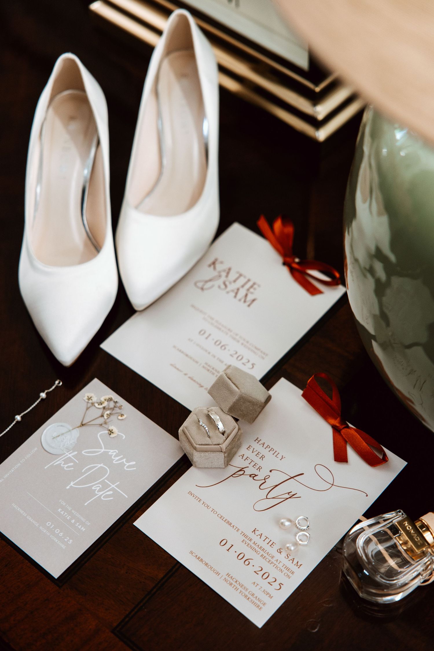 Wedding flat lay featuring white heels, Katie and Sam invitations with red ribbon, ring box, and perfume on dark surface.