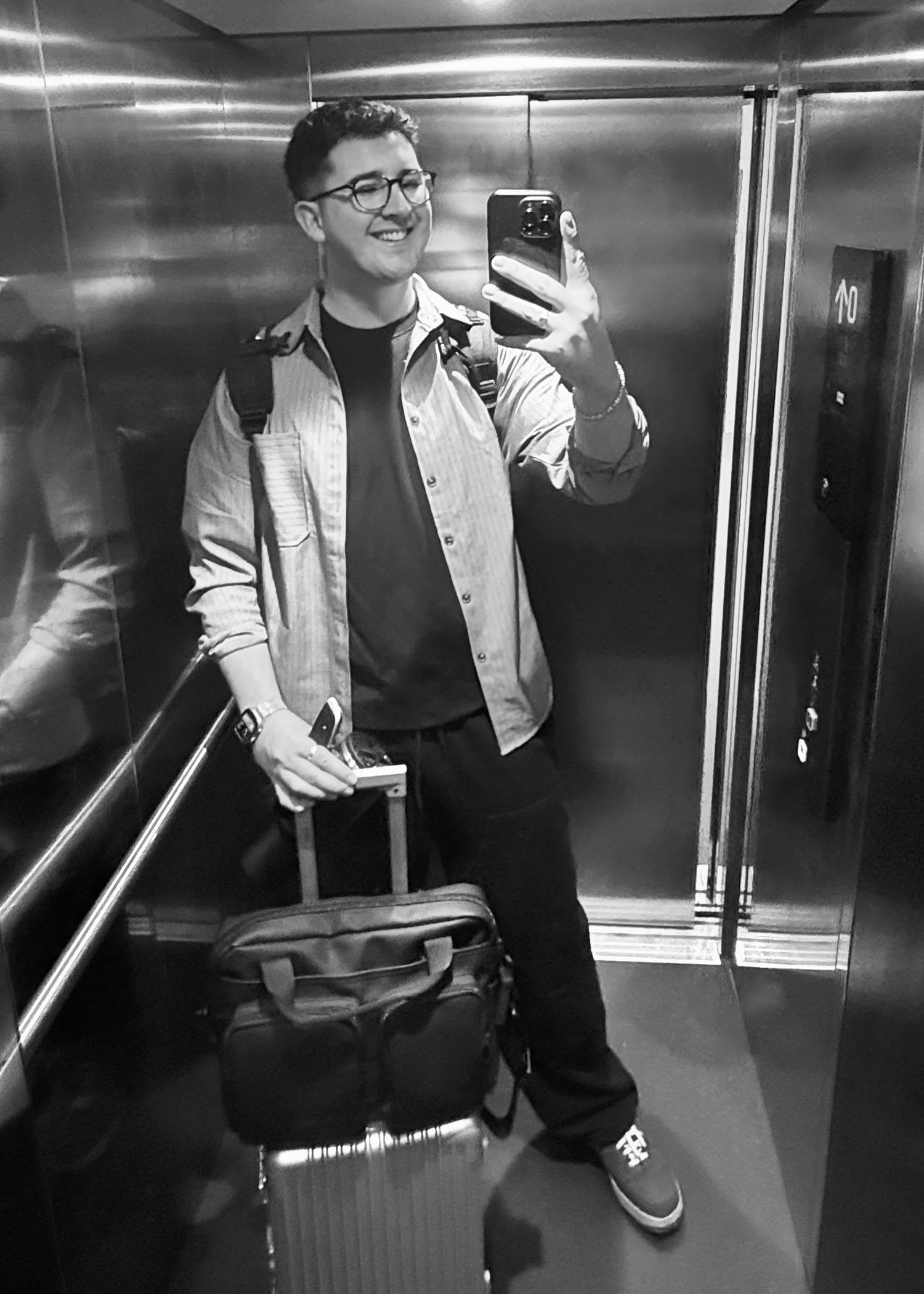 Black and white elevator mirror selfie of smiling man with glasses, luggage and backpack.