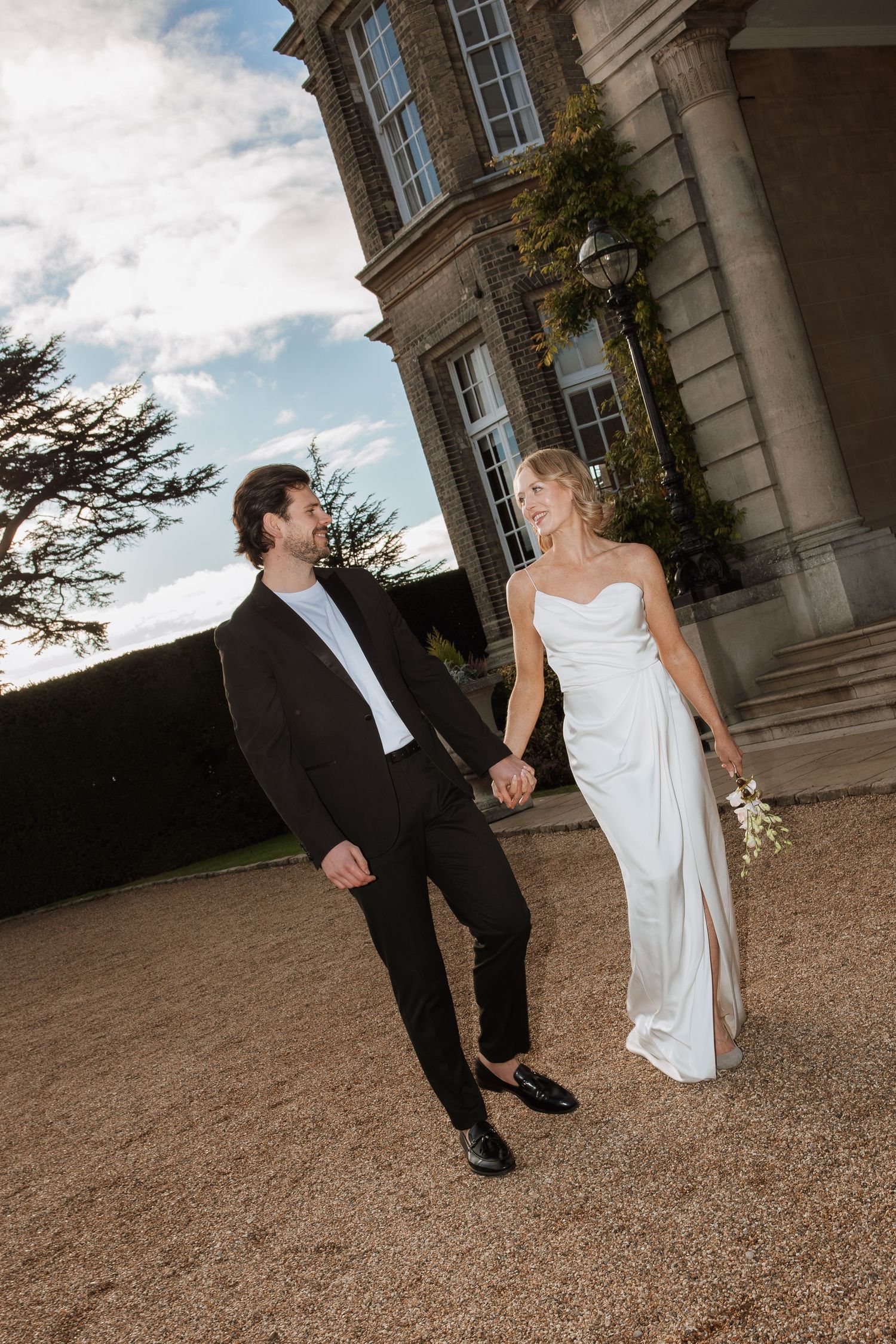 Bride in white strapless wedding dress and groom in black tuxedo walk hand in hand outside elegant estate.