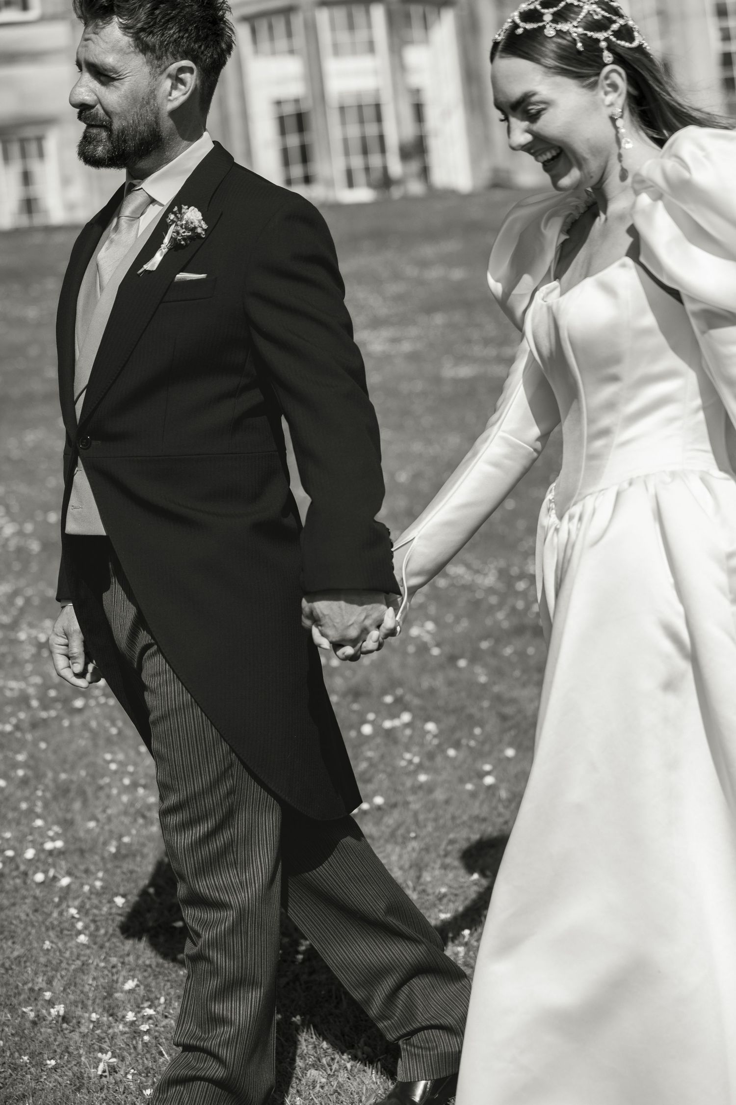 Black and white photo of a smiling bride in white gown and groom in dark suit holding hands walking outdoors.