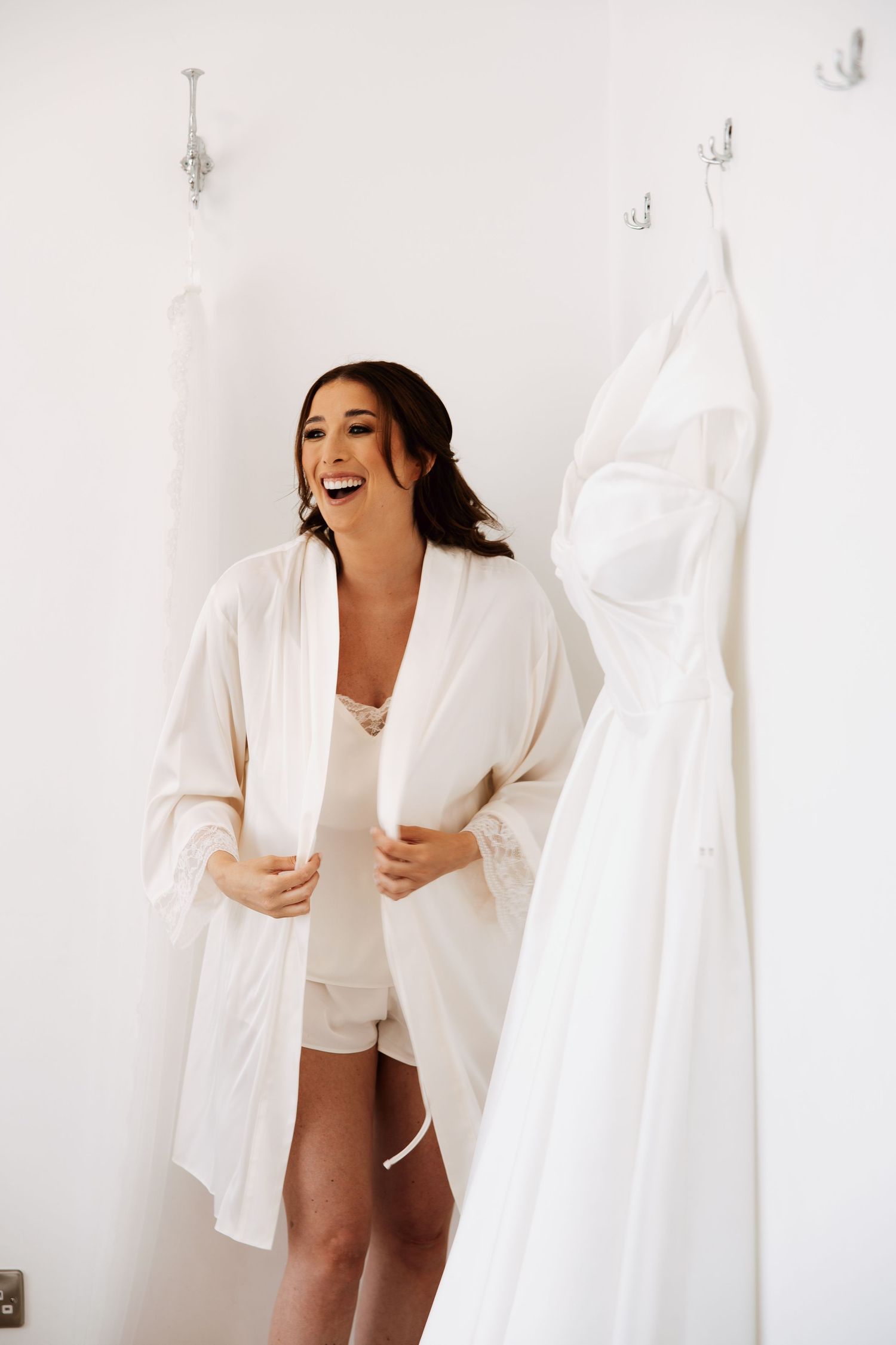 Laughing bride in white robe stands beside her hanging wedding dress in a bright dressing room.