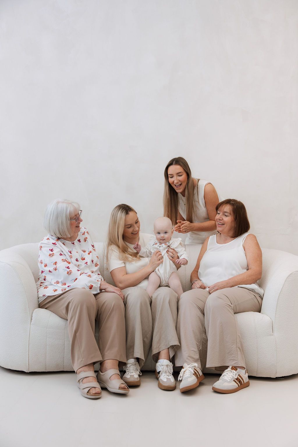 Multi-generational family of women sitting on white sofa admiring newborn baby in bright studio setting.