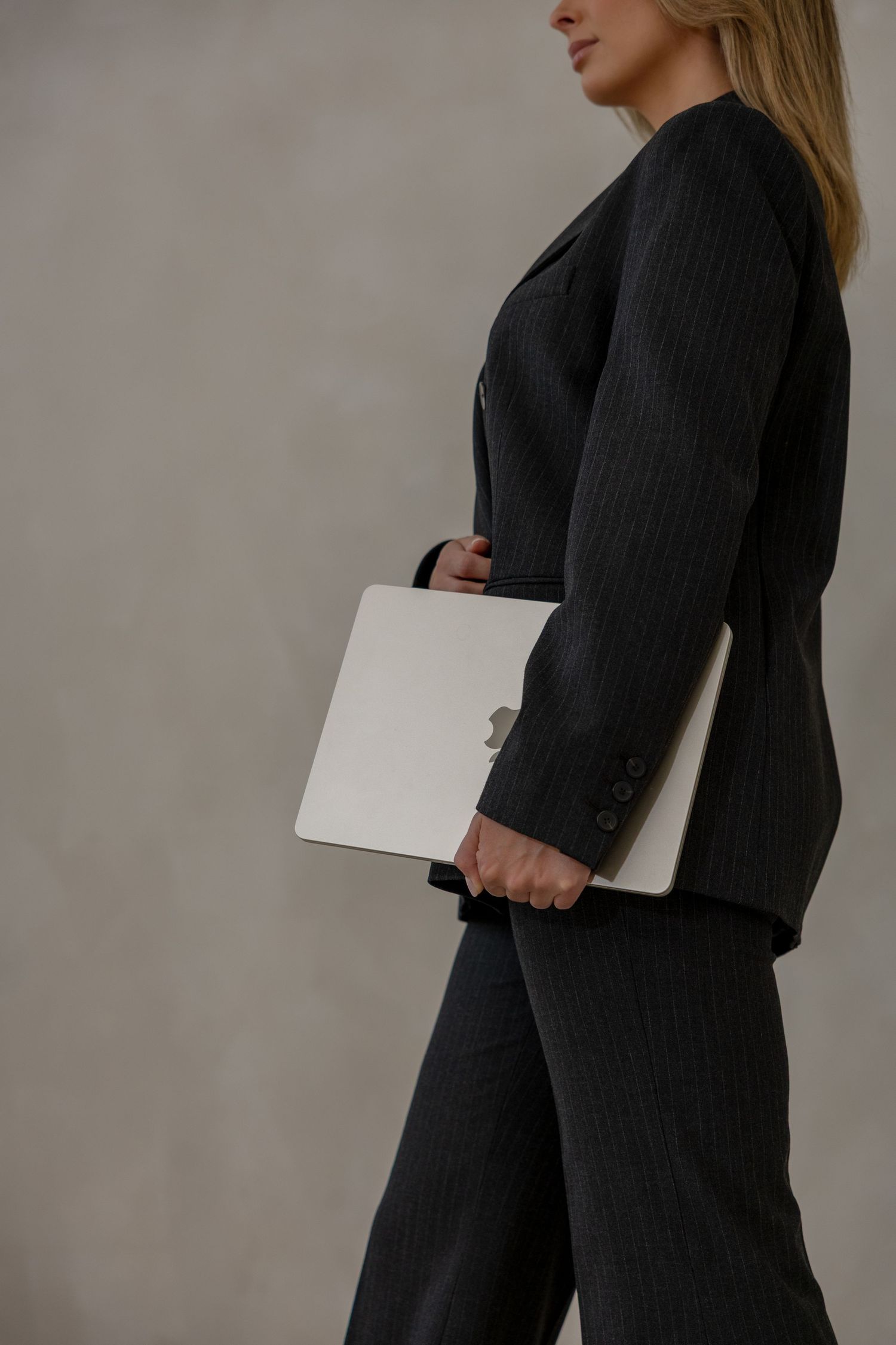 Professional woman in dark pinstripe suit holding a white laptop, standing against a gray background.