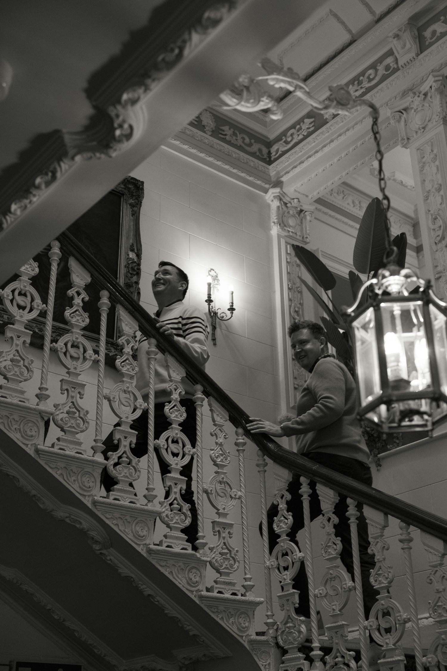 Two men smiling on an ornate staircase with decorative iron railings and a vintage lantern in a grand interior.