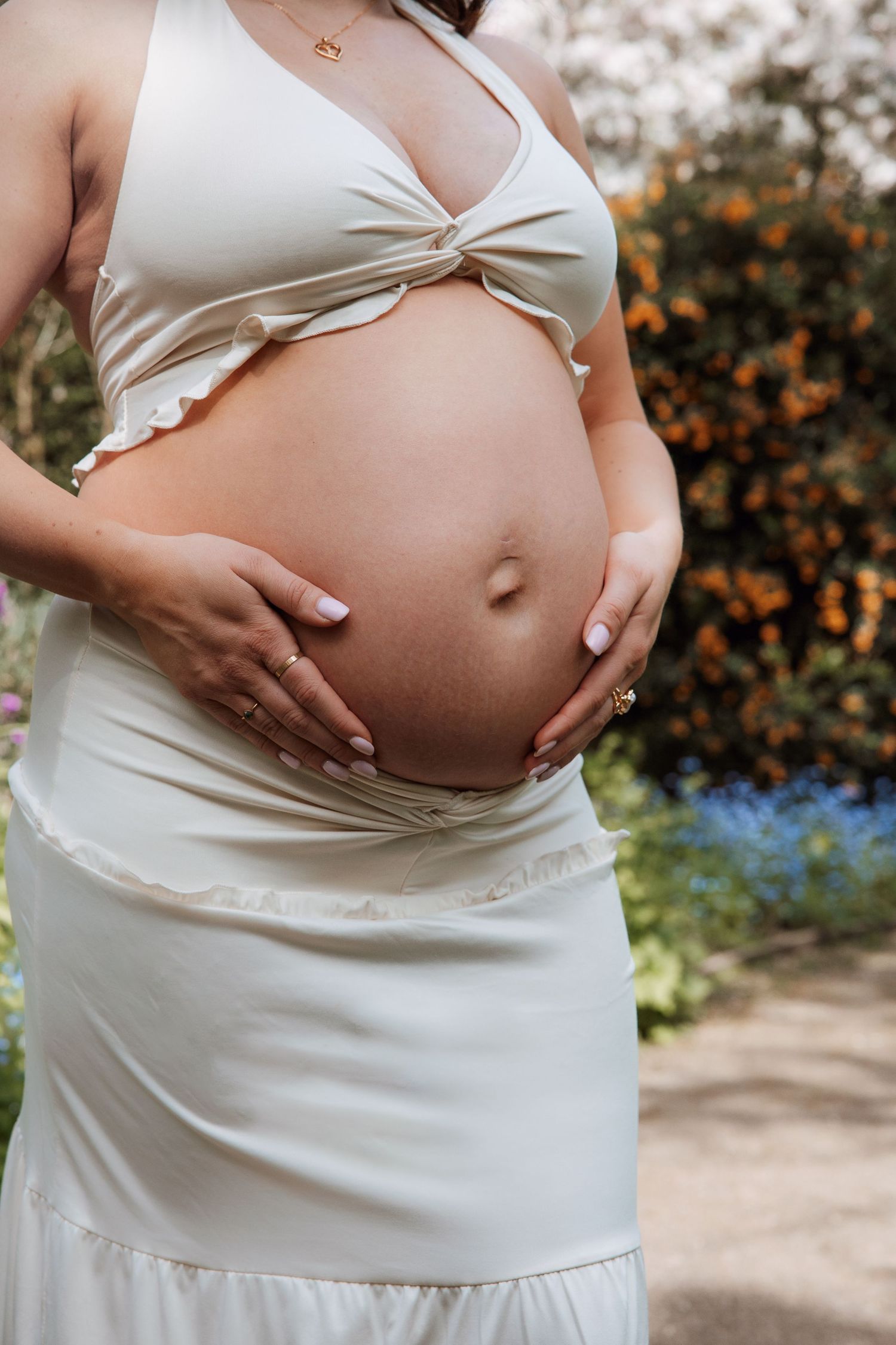 Pregnant woman in white crop top and skirt cradling baby bump outdoors in a garden setting.