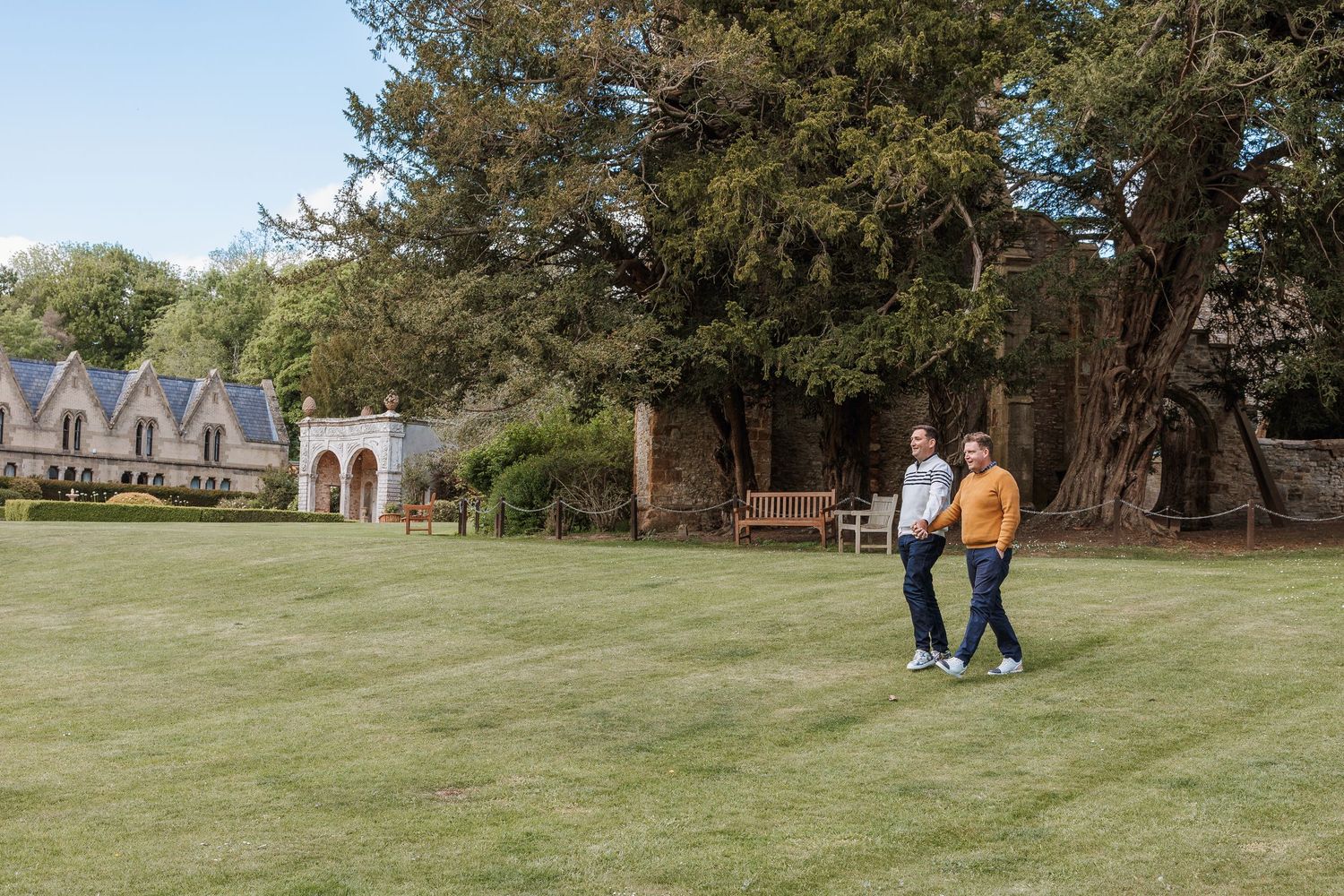 Two people walking on lush green lawn beside historic stone building with large trees on a sunny day.