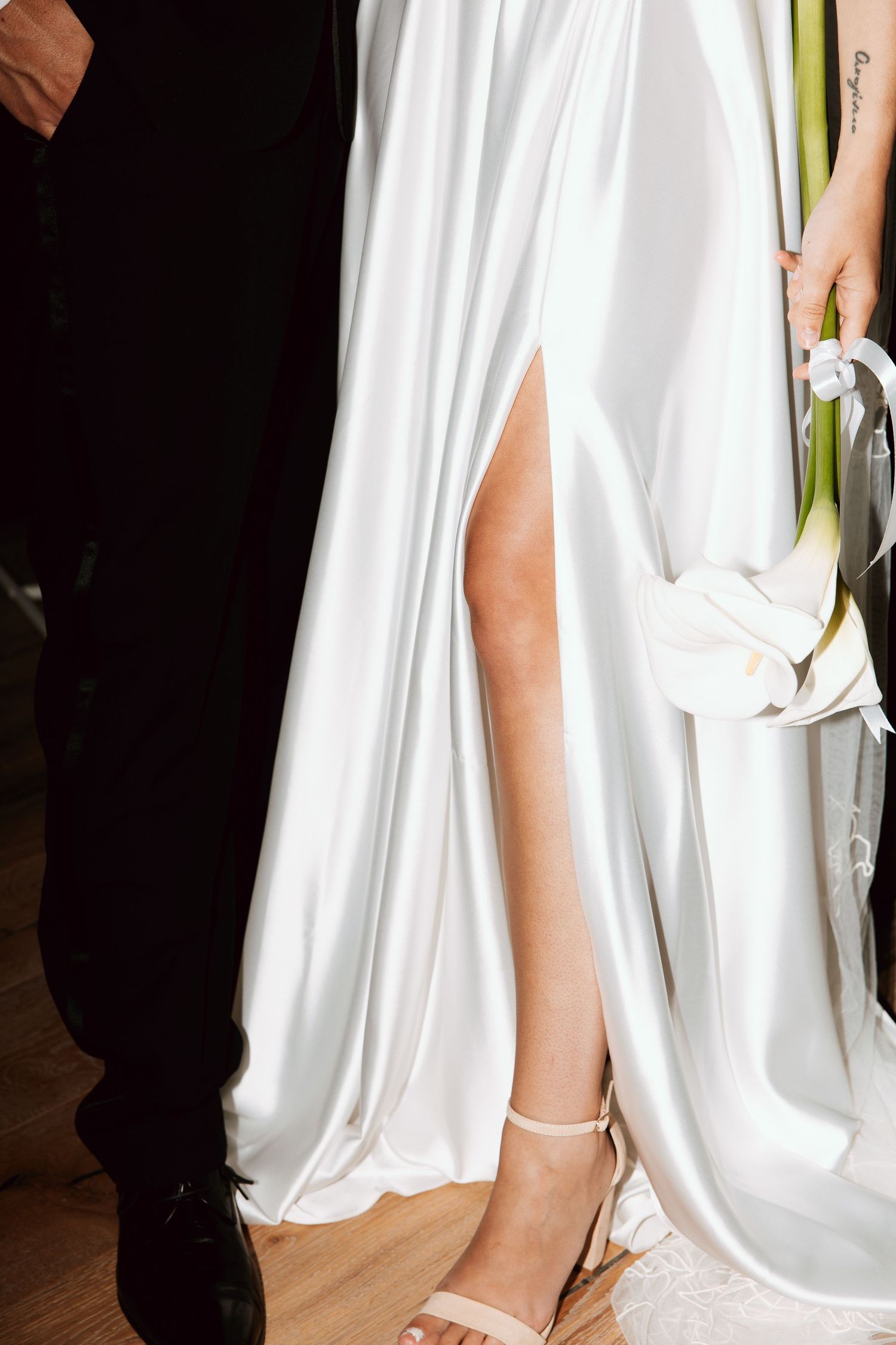 Close-up of a bride in a white satin gown with a thigh-high slit holding a calla lily bouquet.