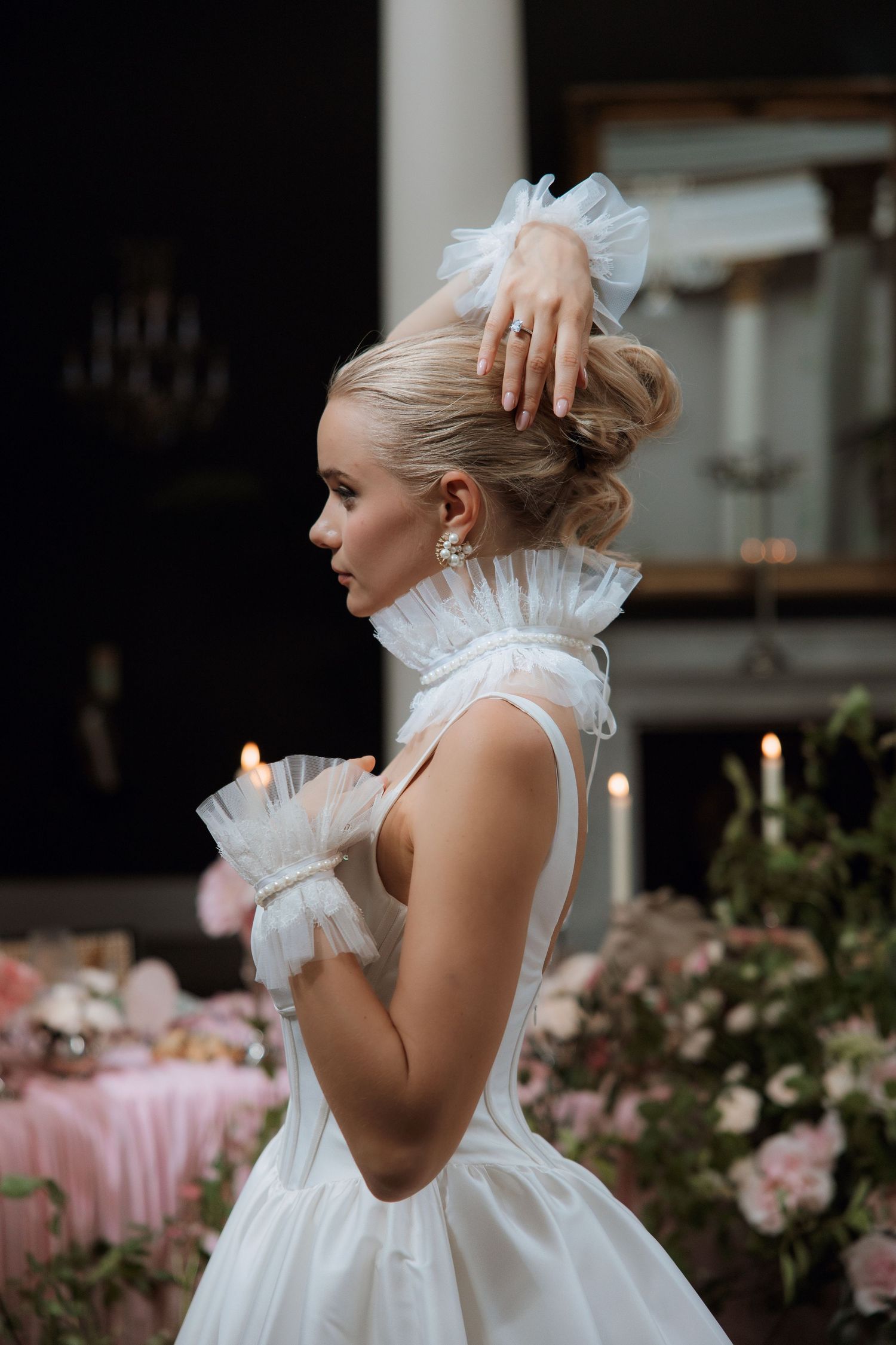 Elegant bride in white ruffled gown with tulle hair bow and updo, posing in a candlelit floral setting.