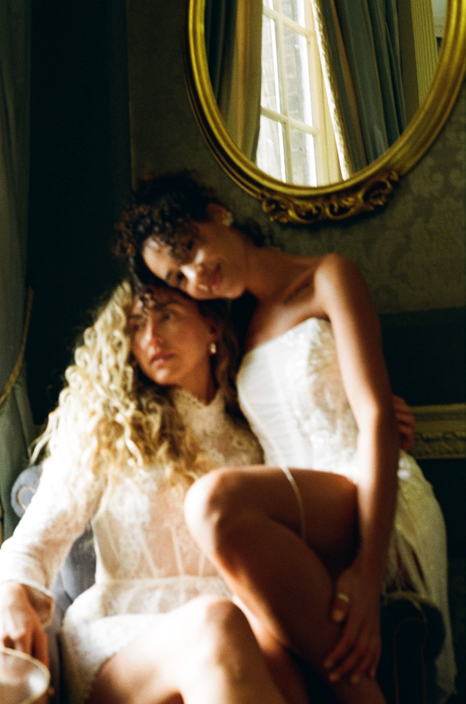 Two women share an intimate embrace in a ornate room, one in white lace bridal lingerie near a gold oval mirror.