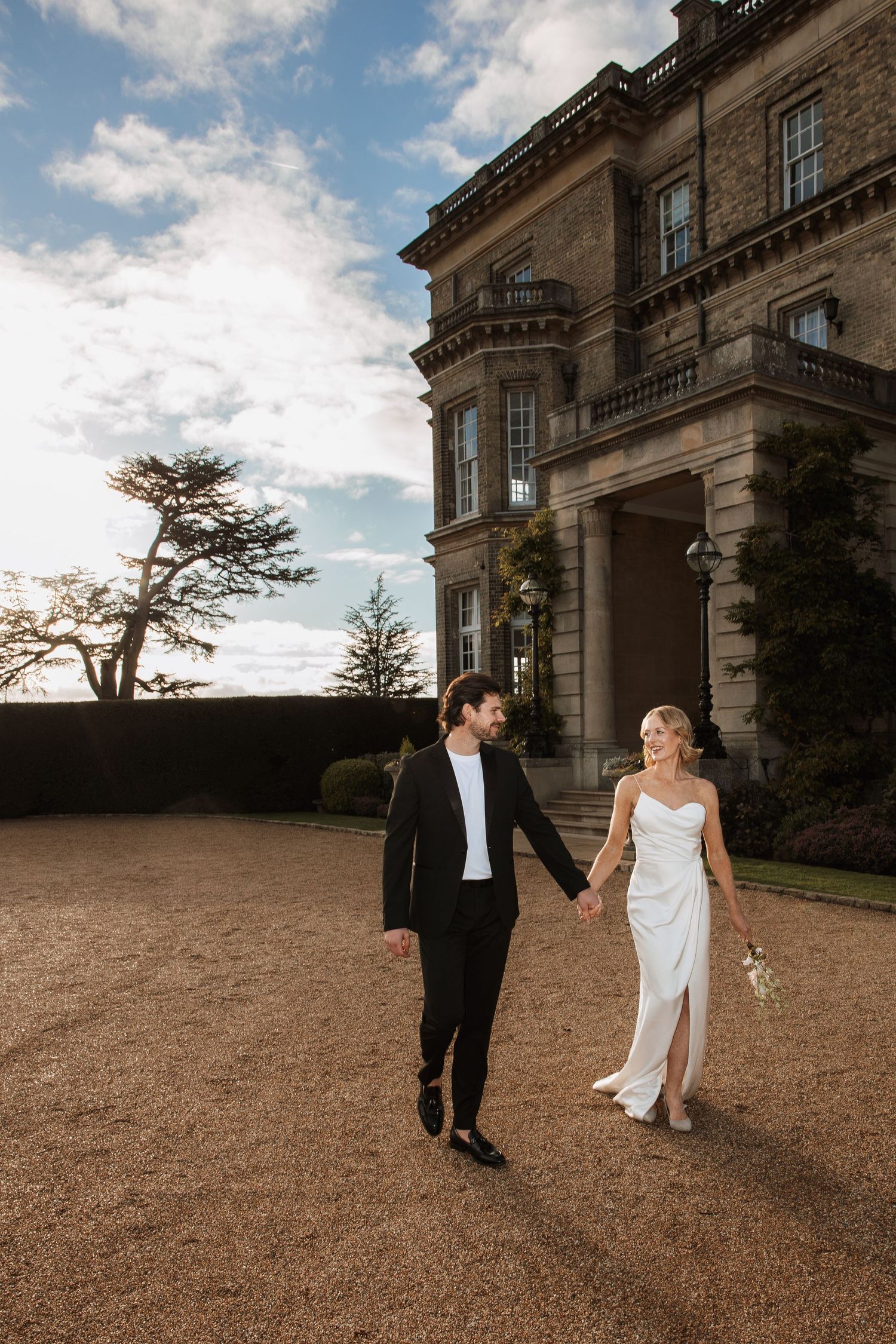 Bride and groom holding hands walking outside a grand stone mansion on their wedding day.