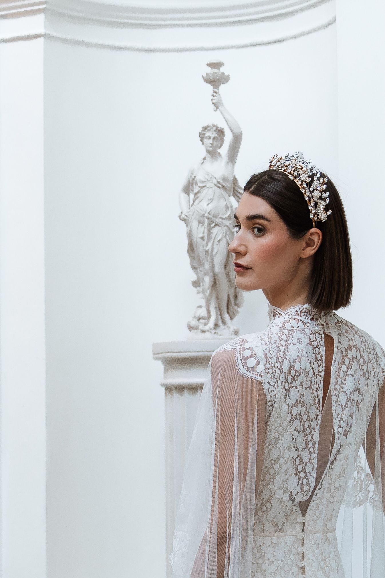 Bride in lace gown with pearl crown poses before classical white statue in elegant architectural setting.