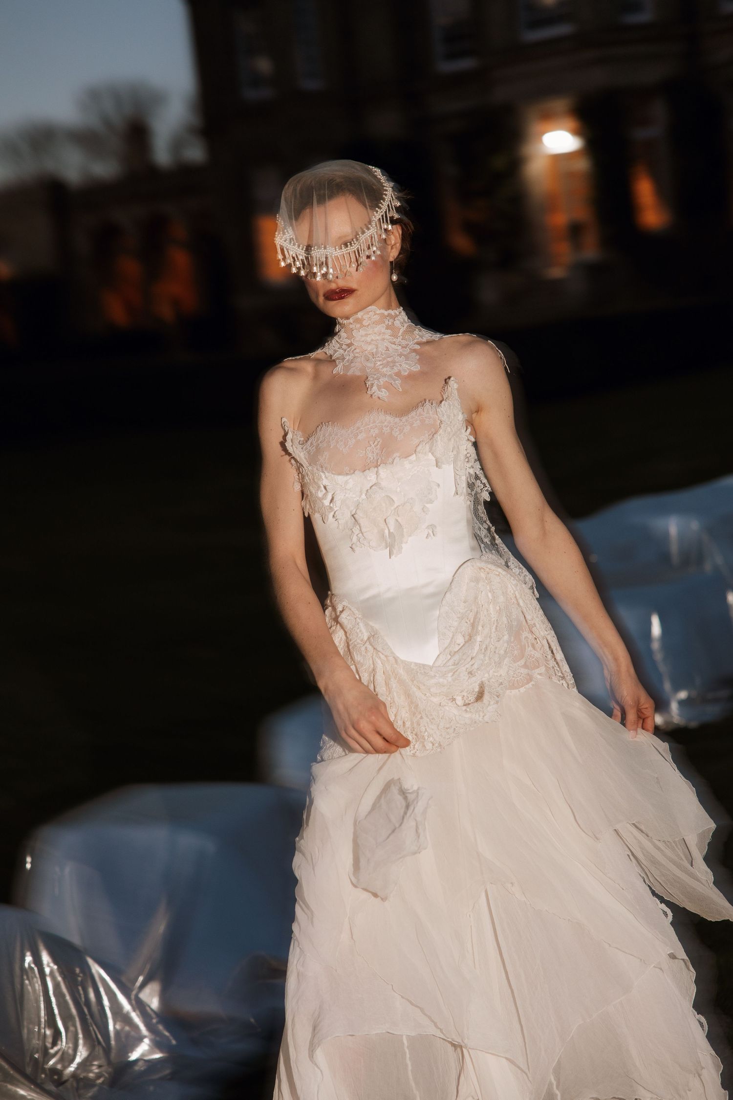 Bride in an elegant lace wedding gown with veil headpiece poses outdoors at dusk near a grand estate.