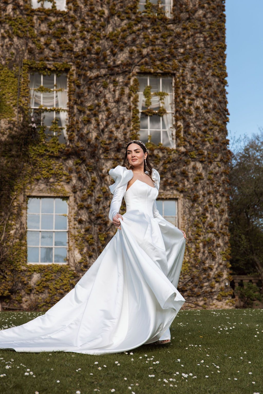 Bride in elegant white satin ball gown with puff sleeves poses on lawn before ivy-covered castle.