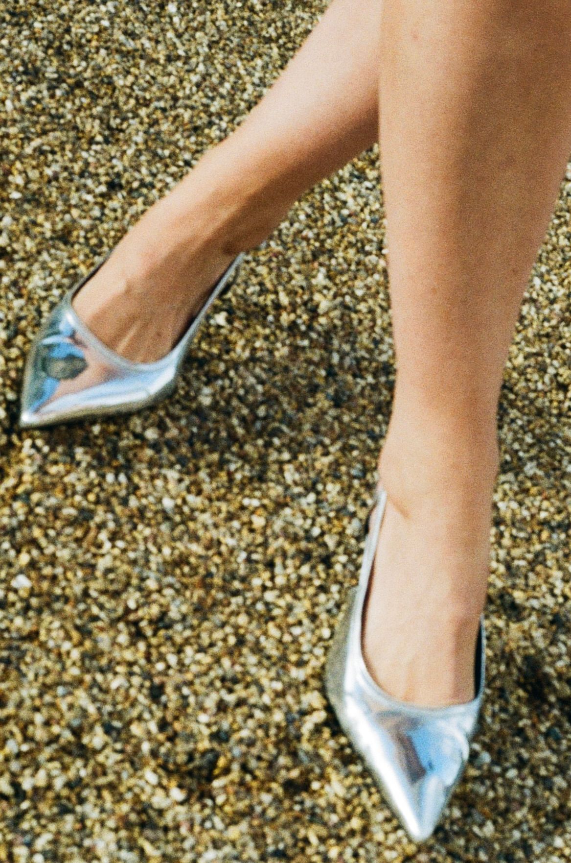 Close-up of bride wearing metallic silver pointed-toe heels on a gravel surface at a wedding.