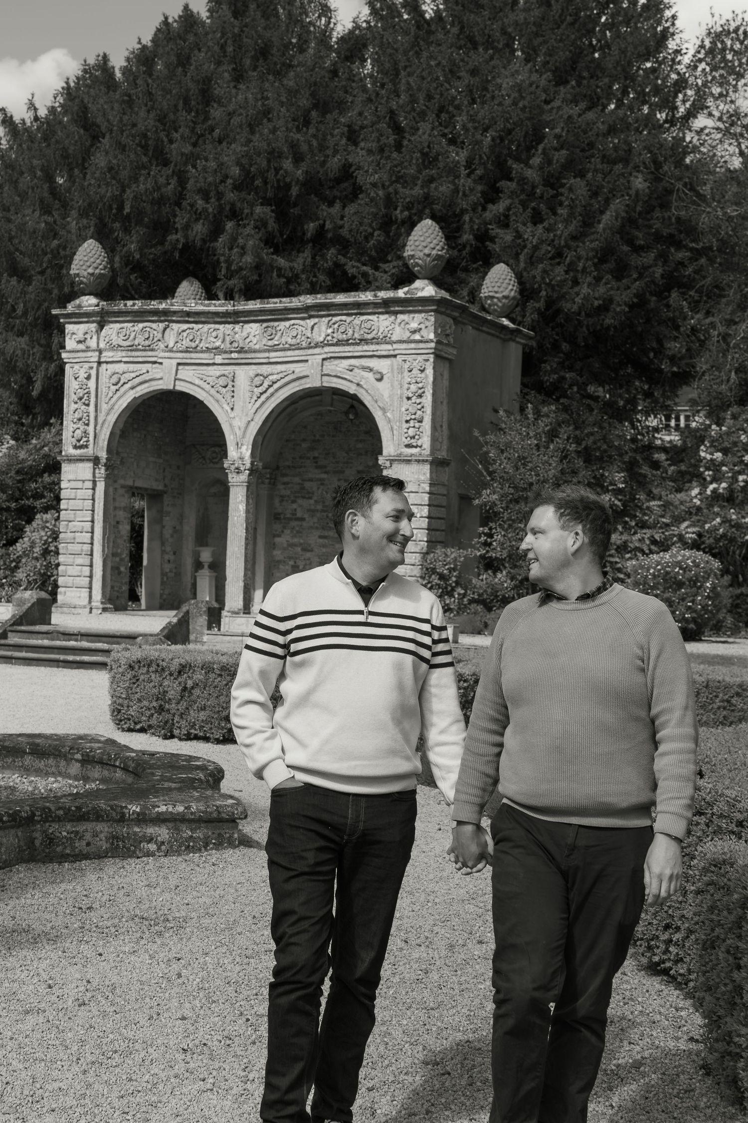 Two men holding hands and smiling while walking in a formal garden with ornate stone archway backdrop.