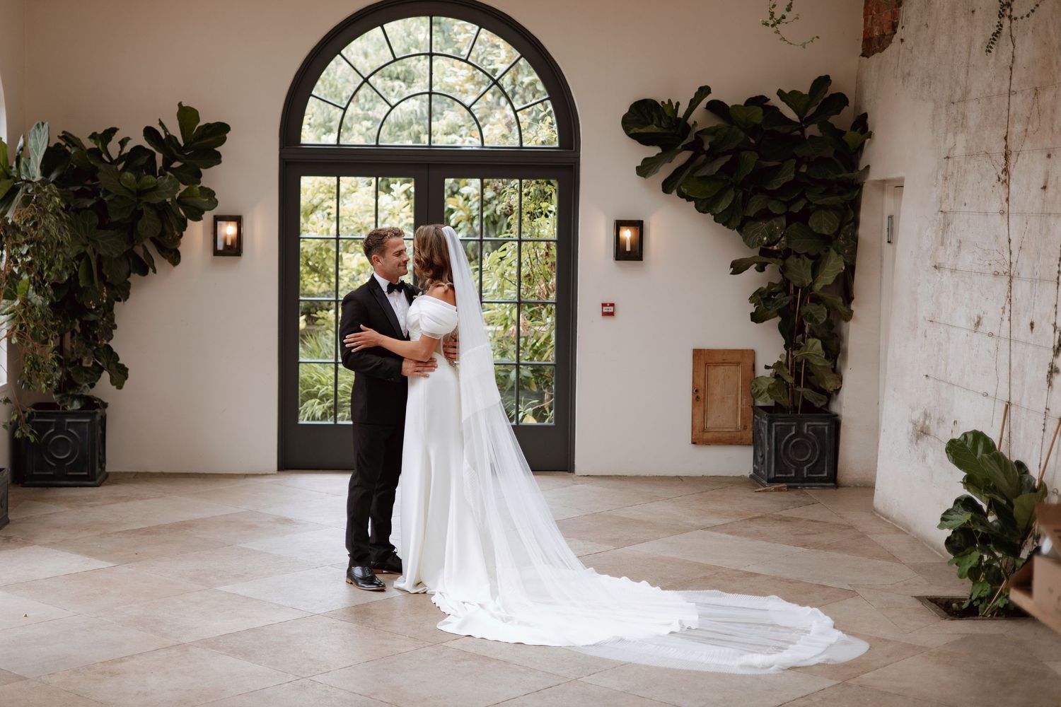 Bride and groom embracing in a bright elegant room with arched French doors and tropical greenery.