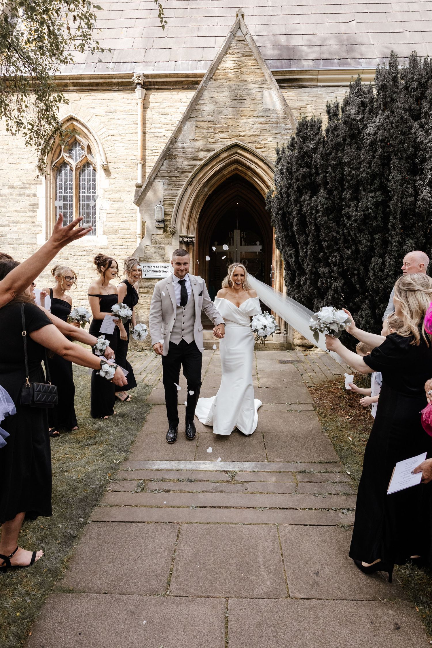 Bride and groom exit stone church while guests throw confetti, bridesmaids in black dresses lining the path.