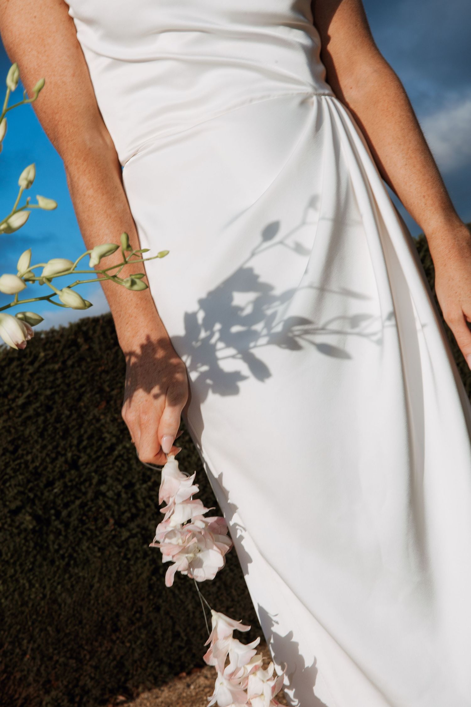 Close-up of a bride in a white satin wedding dress holding delicate white flowers outdoors.
