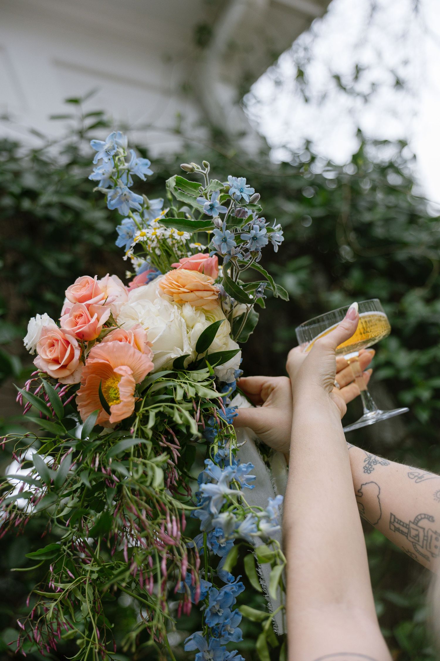 A tattooed hand raises a champagne glass beside a lush floral arrangement of pink roses, blue delphiniums, and greenery outdoors.