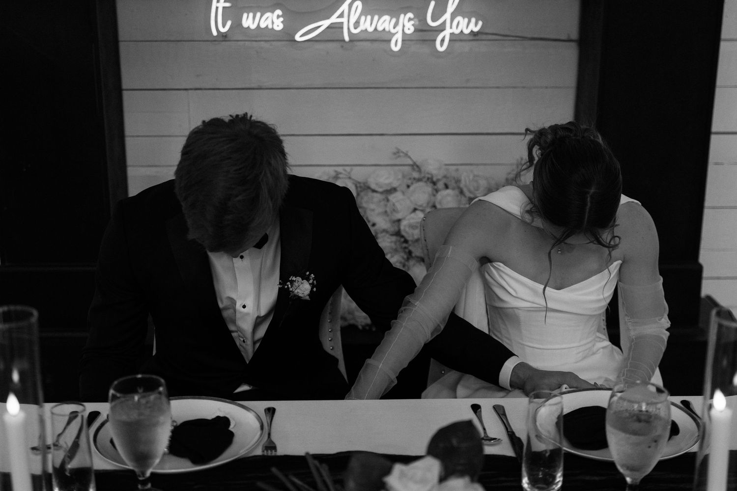 Black and white photo of bride and groom at sweetheart table under It Was Always You neon sign.