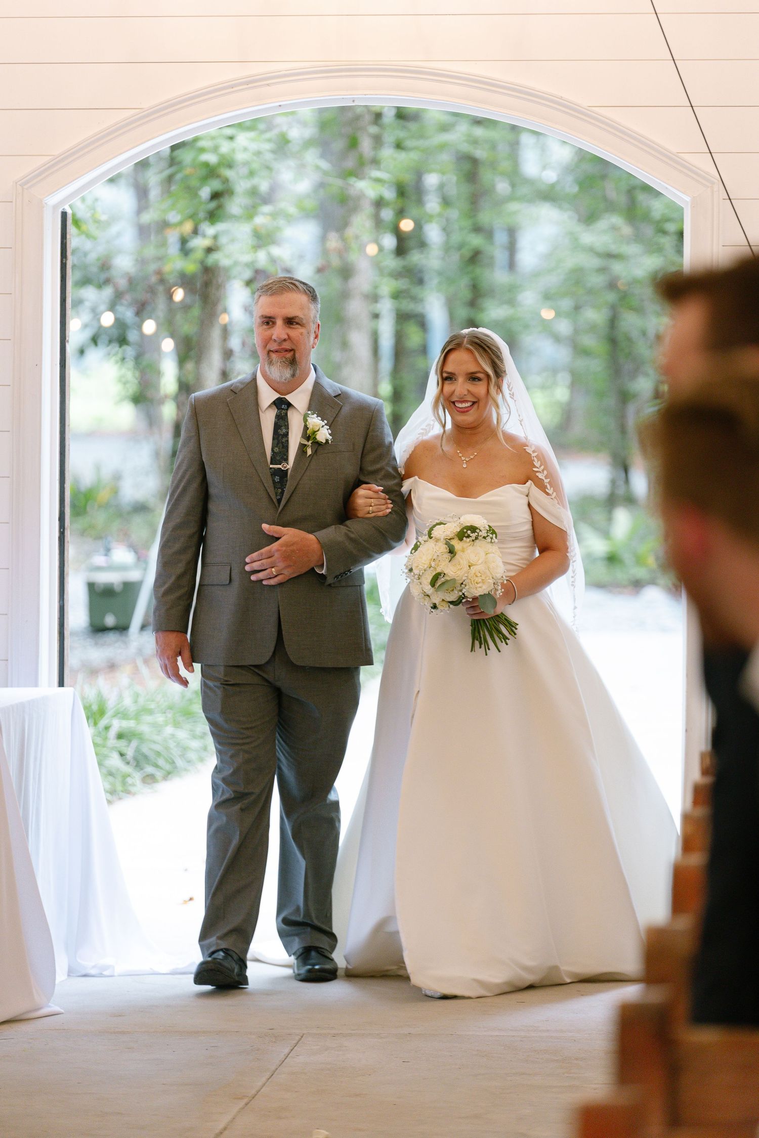 Bride walks down the aisle with her father in a white gown and veil, holding a white bouquet at a wedding ceremony.