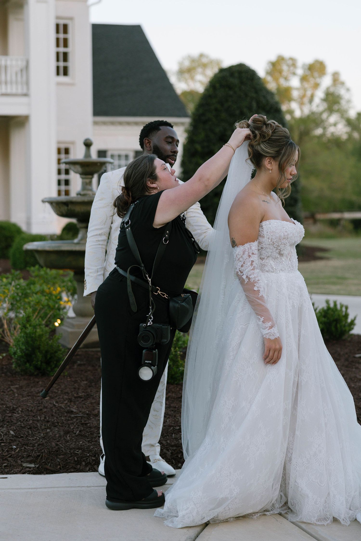 A wedding coordinator adjusts the bride's veil outdoors near a elegant white manor while the groom looks on.