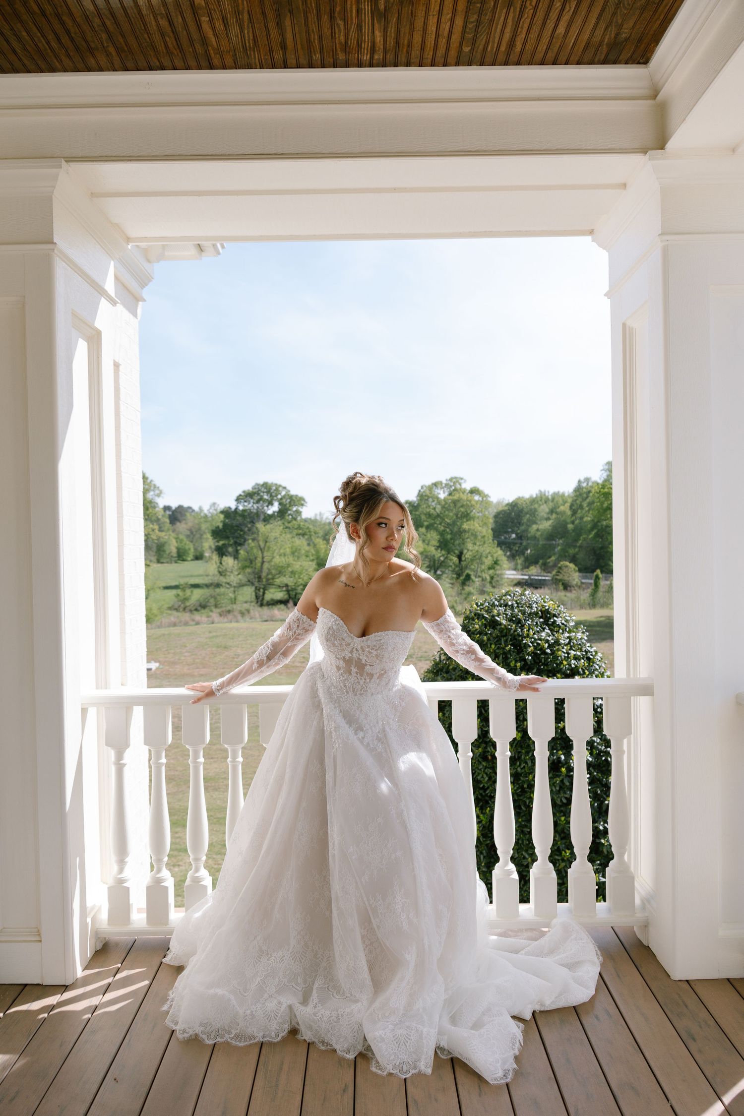 Bride in off-shoulder lace ball gown stands on white balcony railing, smiling with green landscape behind her.