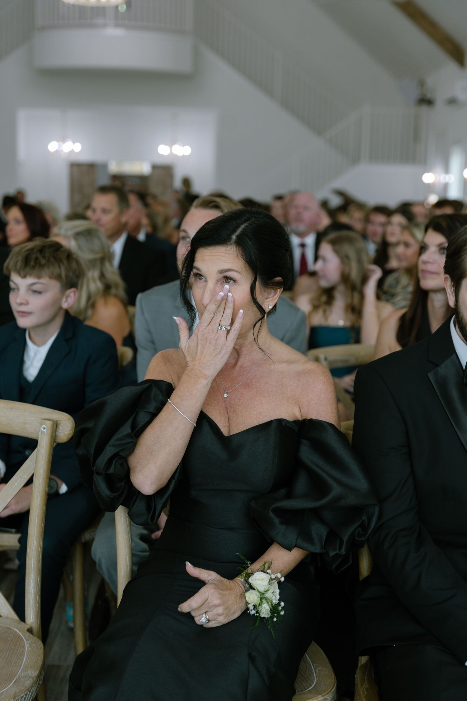 Emotional mother of bride crying in black off-shoulder gown during wedding ceremony in white chapel.