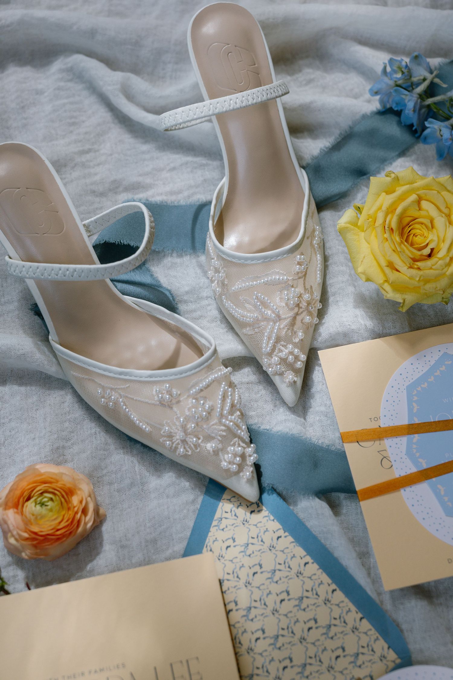 Elegant beaded white mule heels with ankle strap displayed with yellow rose, ranunculus, and blue hydrangeas on soft fabric.