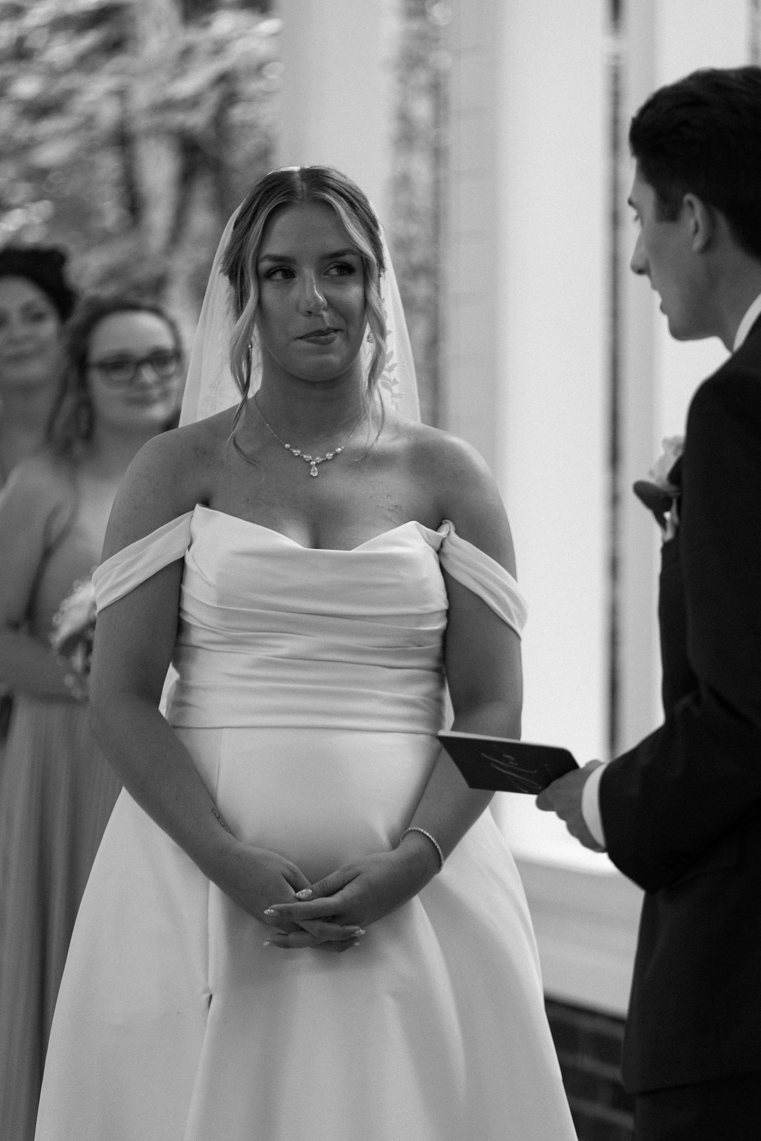 Beautiful bride in off-shoulder white gown and veil reciting vows at outdoor wedding ceremony.